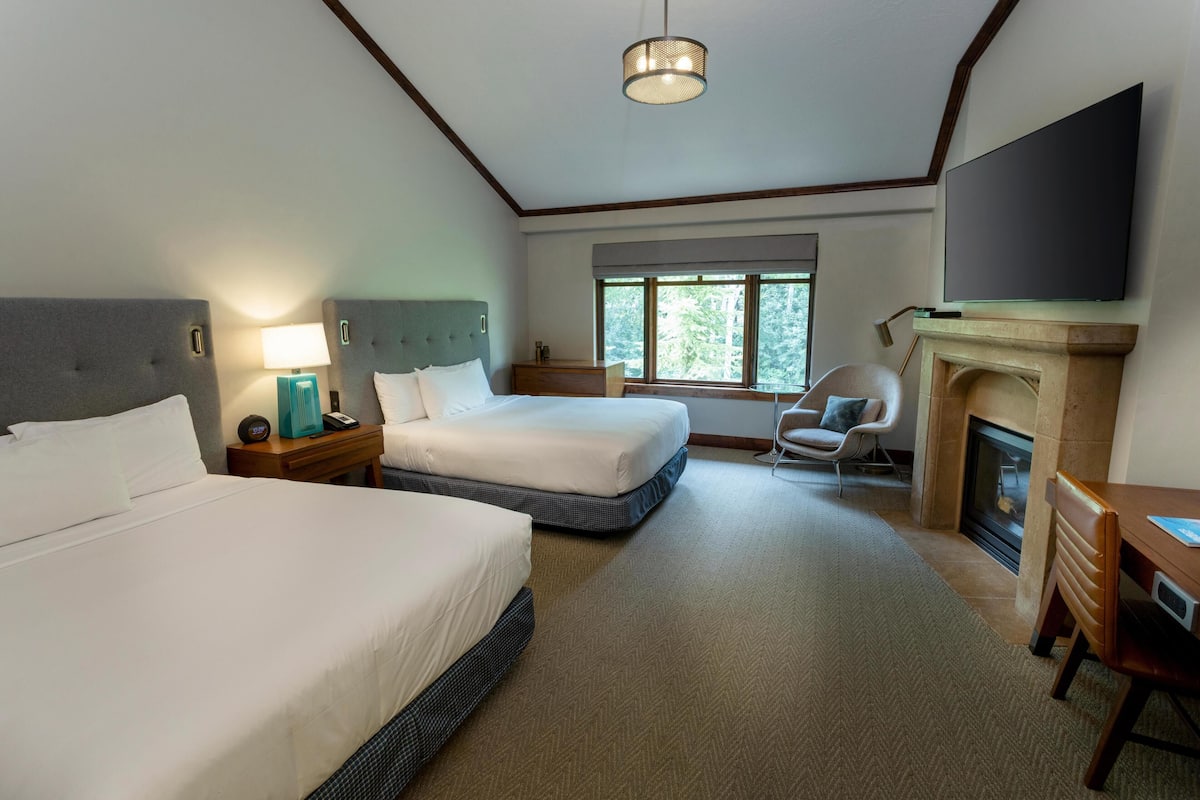 A hotel room with two beds, a television, a fireplace, and a window with a view of trees.