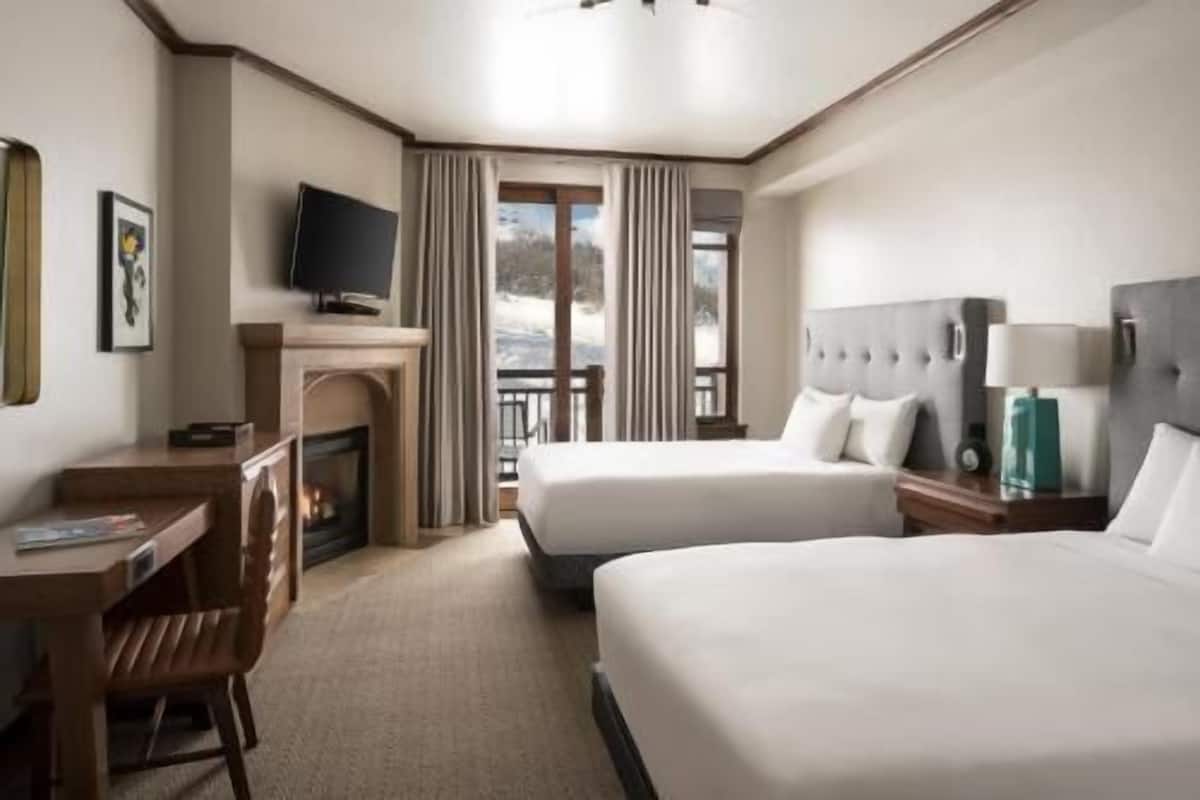 A hotel room with two beds, a desk, a television, and a fireplace.