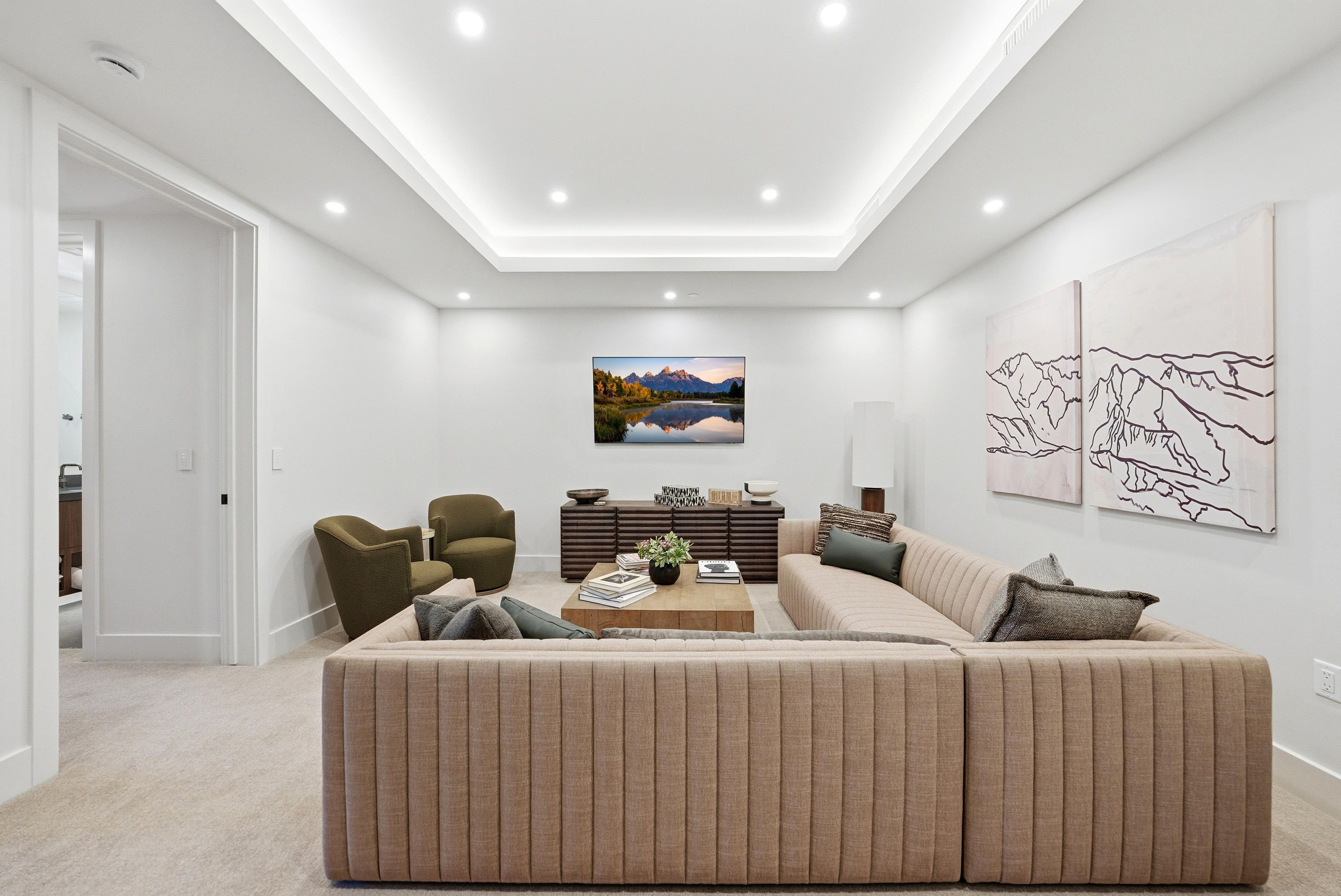 A modern living room with a large beige sectional sofa, a wooden coffee table with books and a plant, a dark wood sideboard, and a wall-mounted TV.