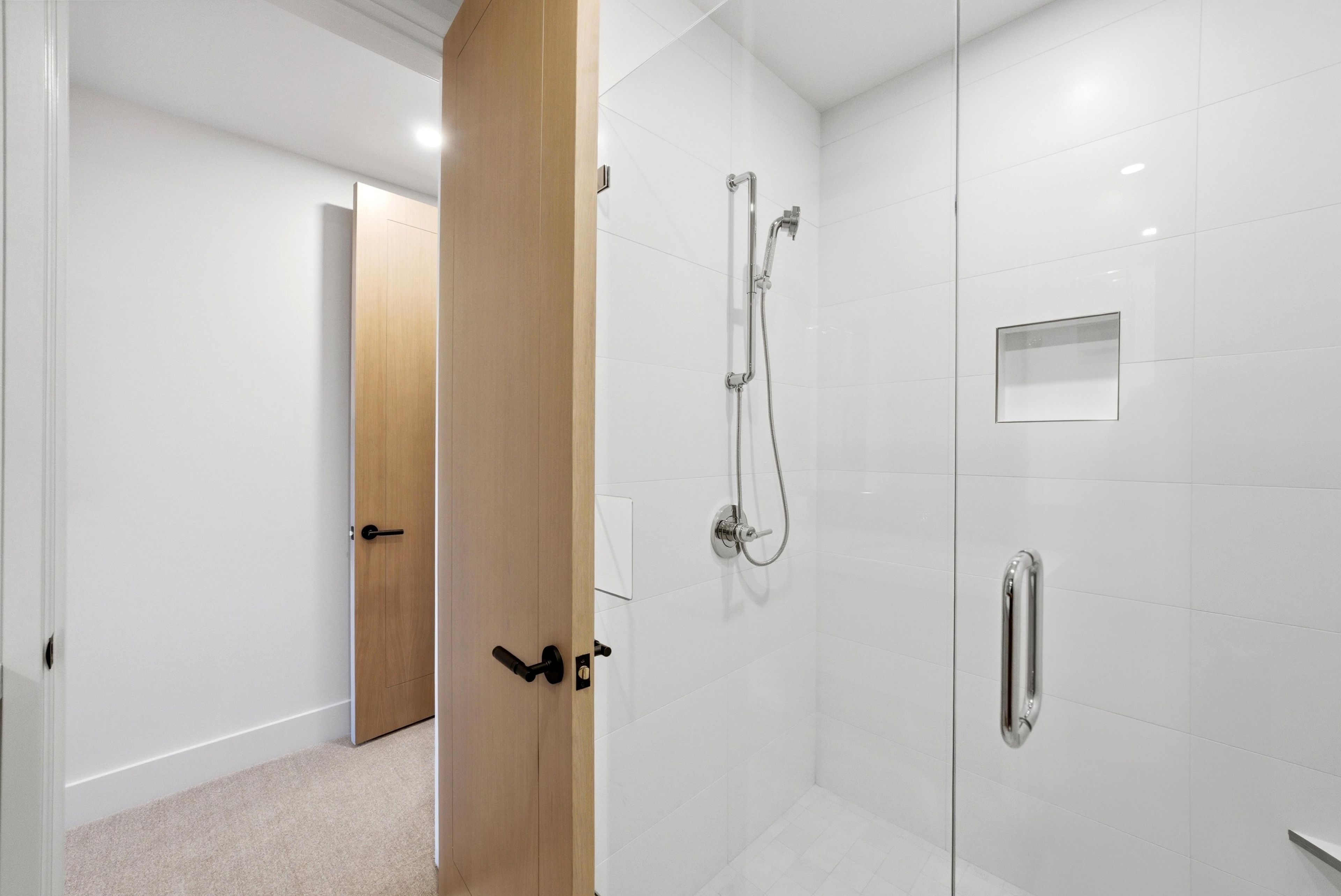 Un baño moderno con cabina de ducha de vidrio, puerta de madera y pared revestida de azulejos blancos.