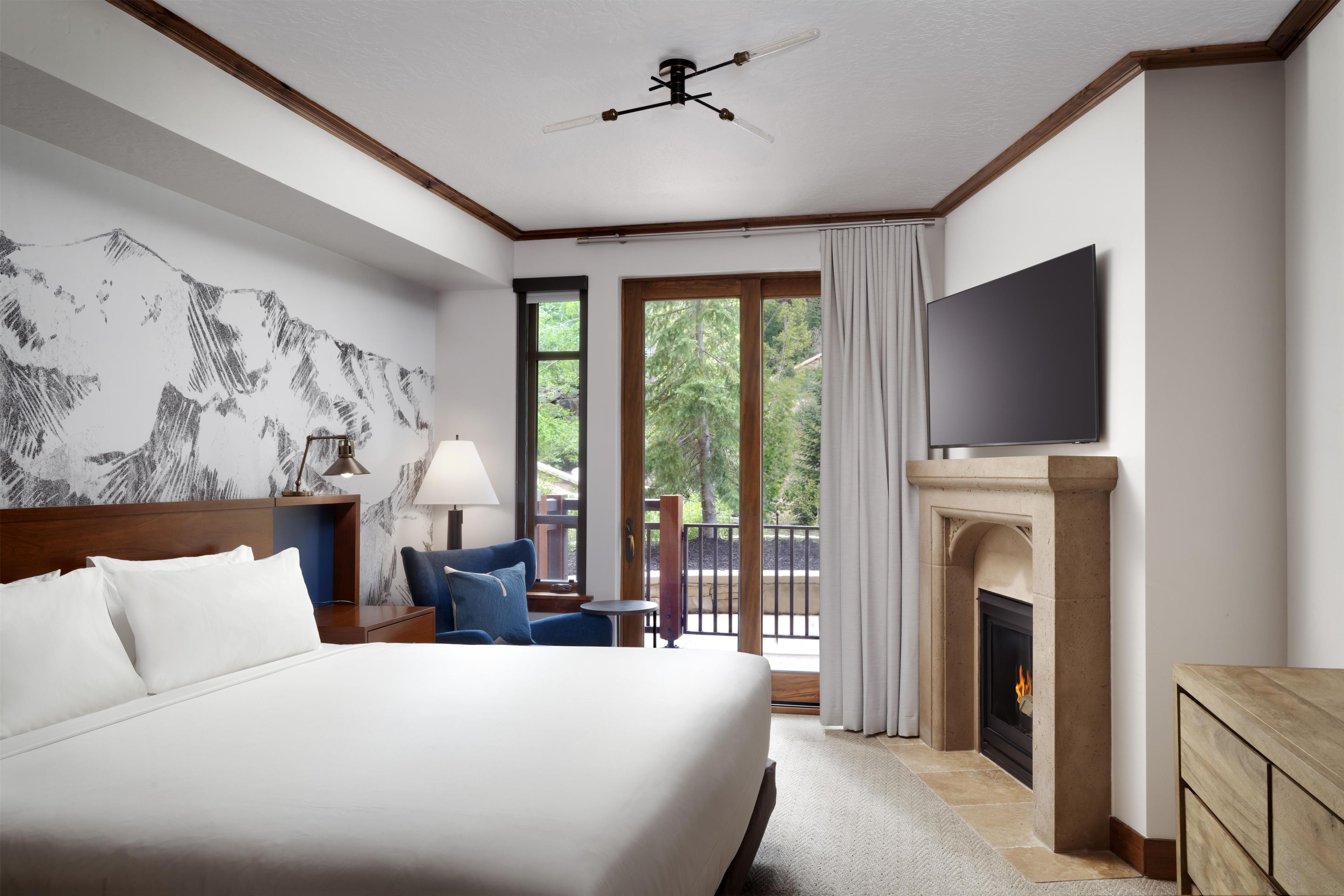 A hotel room with a large bed, a flat-screen TV, a fireplace, and a balcony with a view of trees.