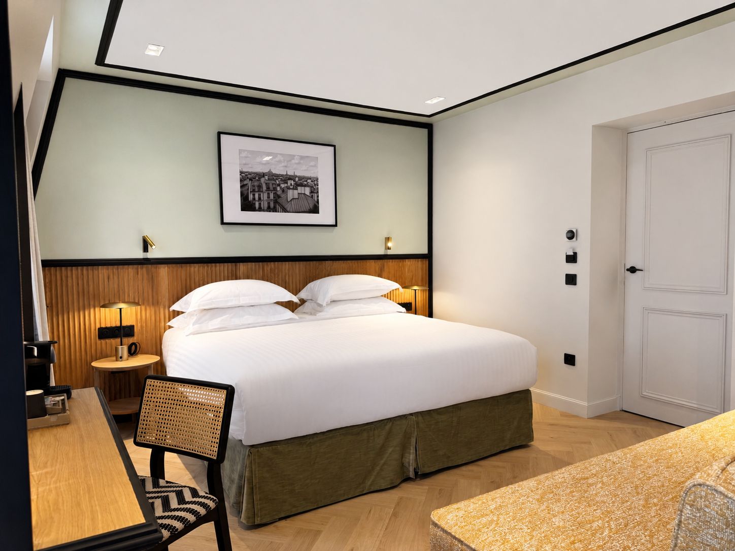 A hotel room with a large bed, a desk, a chair, a bedside table, a lamp, a framed picture on the wall, and a door.