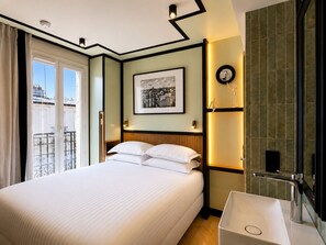 A hotel room with a bed, a bedside table, a mirror, and a view of the outside.