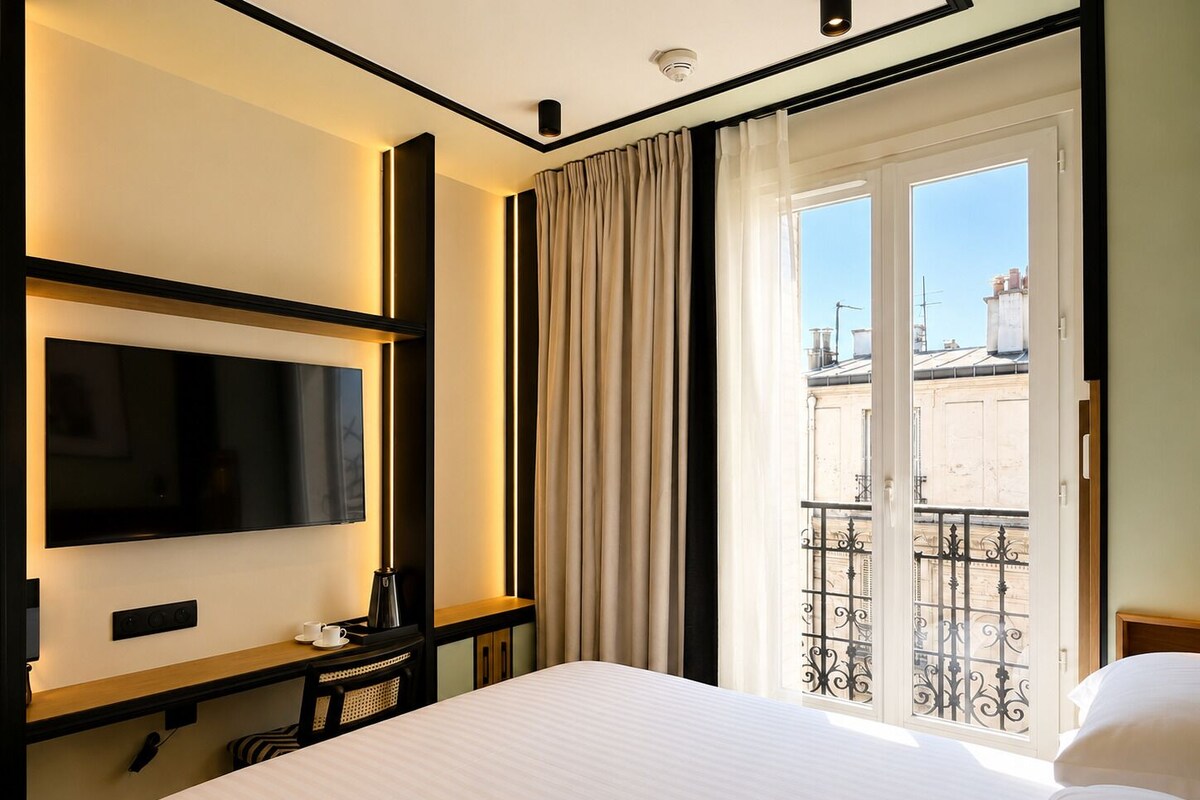 A hotel room with a bed, a flat-screen TV mounted on the wall, a small table with a cup and a remote control, and a window with curtains.