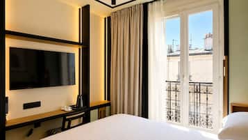 Une chambre d’hôtel avec un lit, un téléviseur à écran plat fixé au mur, une petite table avec une tasse et une télécommande, et une fenêtre avec des rideaux.