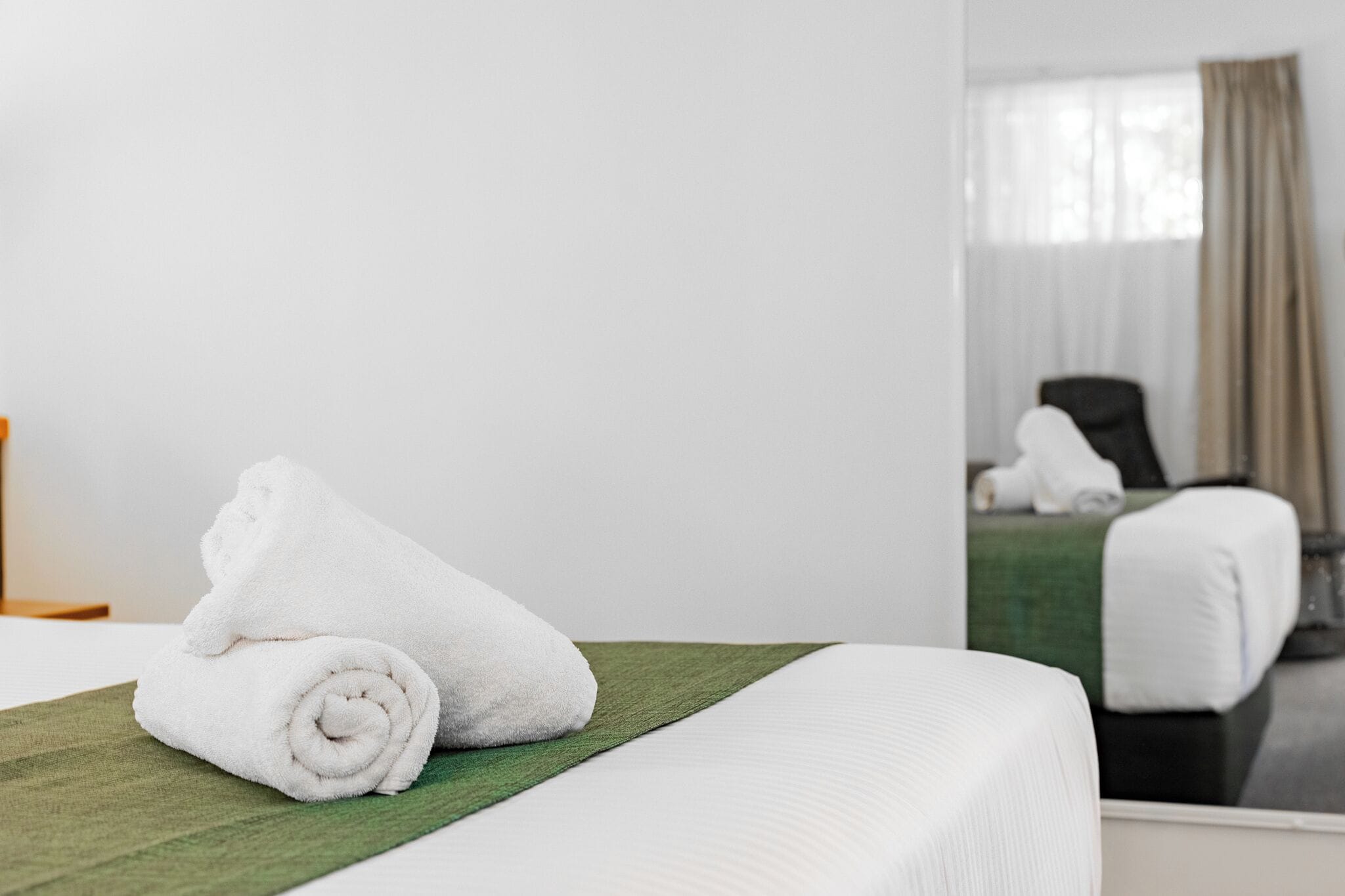 A neatly made bed with white towels, a green bedspread, and a mirror reflecting a room with a chair and a window with curtains.