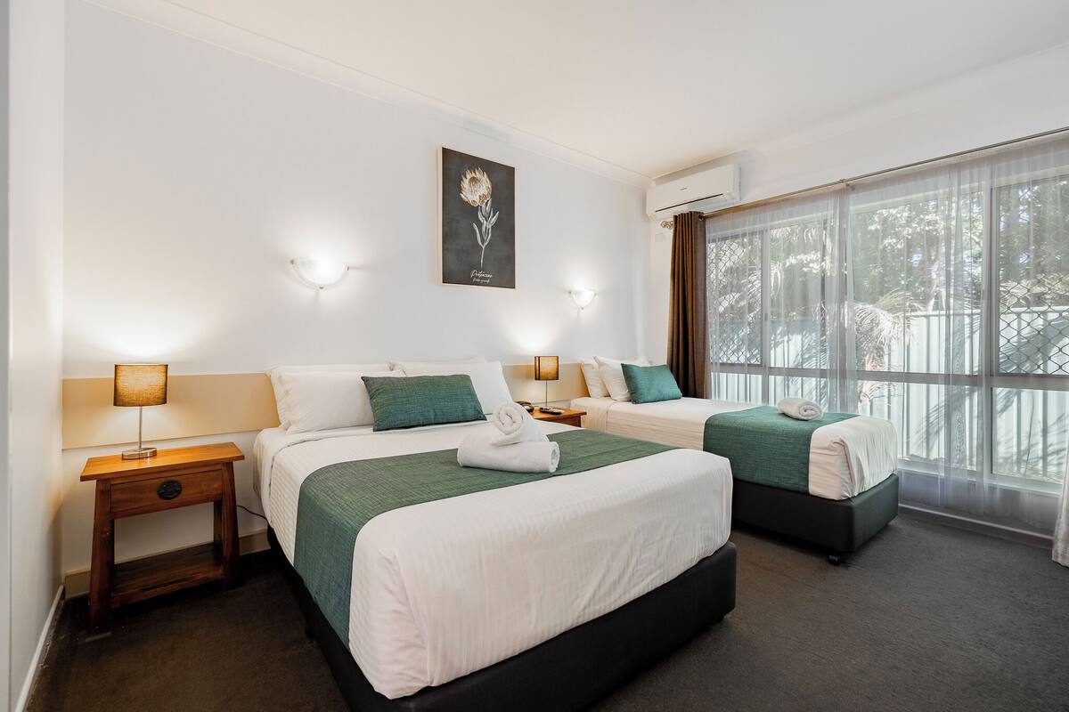 A hotel room with two beds, a large window with curtains, a wall-mounted artwork, and bedside lamps.