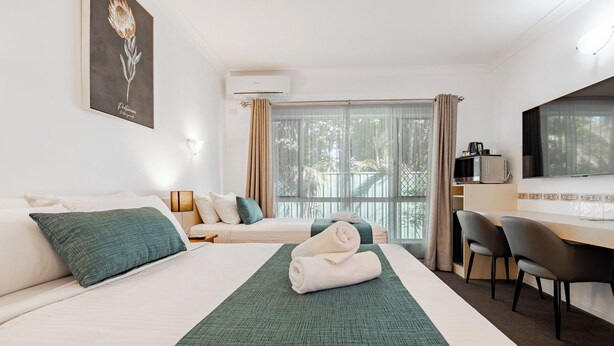 A hotel room with a bed, a desk, and a chair. There is a window with curtains, a wall-mounted air conditioner, and a framed picture on the wall.