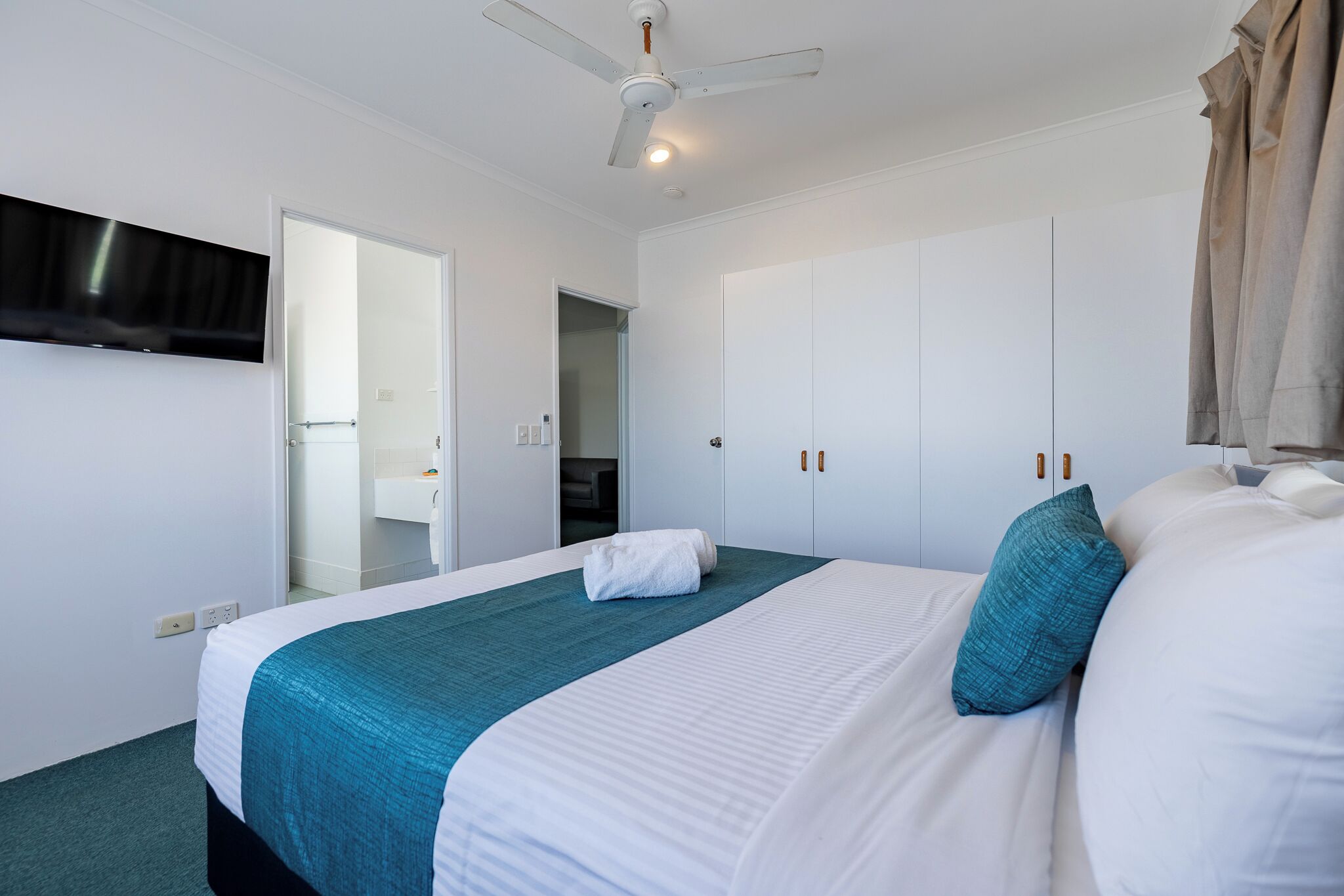 Una habitación de hotel con cama, ventilador de techo, televisor montado en la pared y armario.