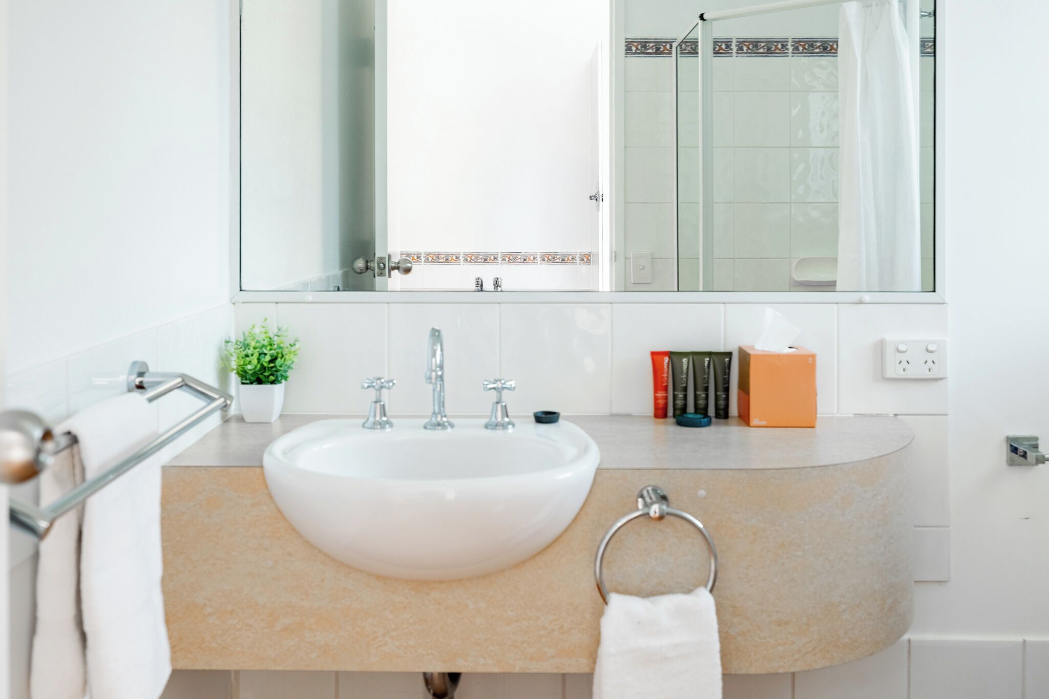 Een badkamer met een wit wastafelmeubel, een spiegel, een handdoekhouder, een kleine potplant en toiletartikelen op het aanrecht.