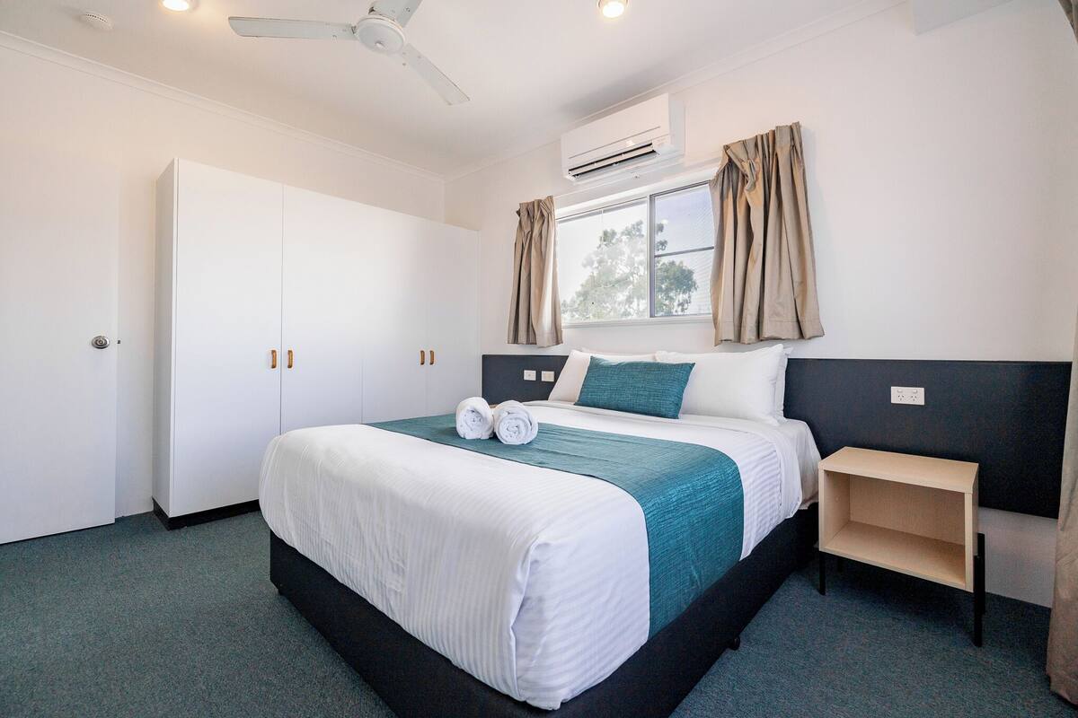 A hotel room with a bed, a nightstand, a wardrobe, and an air conditioning unit.
