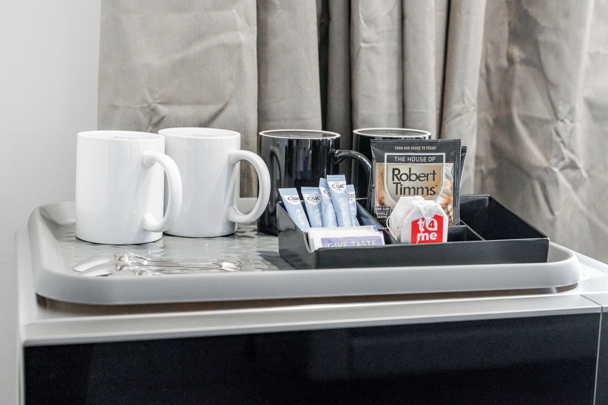 A tray with two white mugs, a black mug, and a box containing a magazine titled 'The House of Robert Timms' along with some packets.