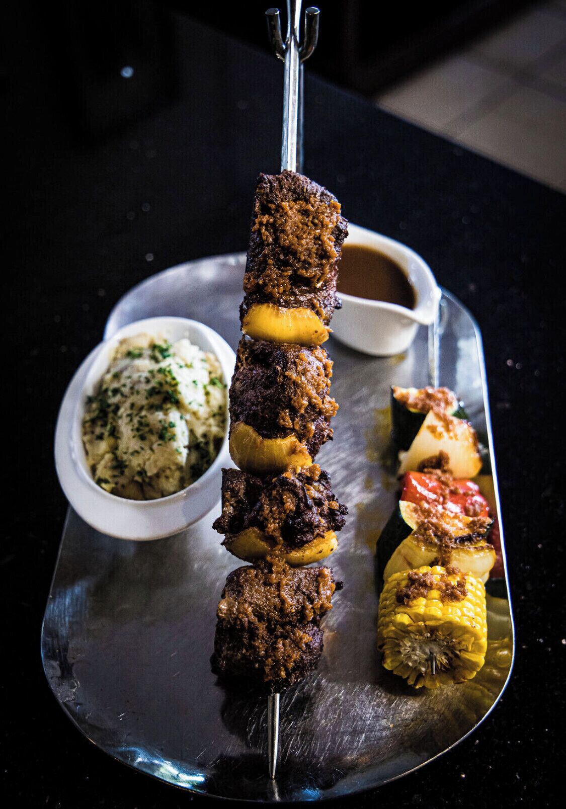 A skewered meat dish with vegetables on a metal tray, accompanied by a bowl of sauce and a side of mashed potatoes.