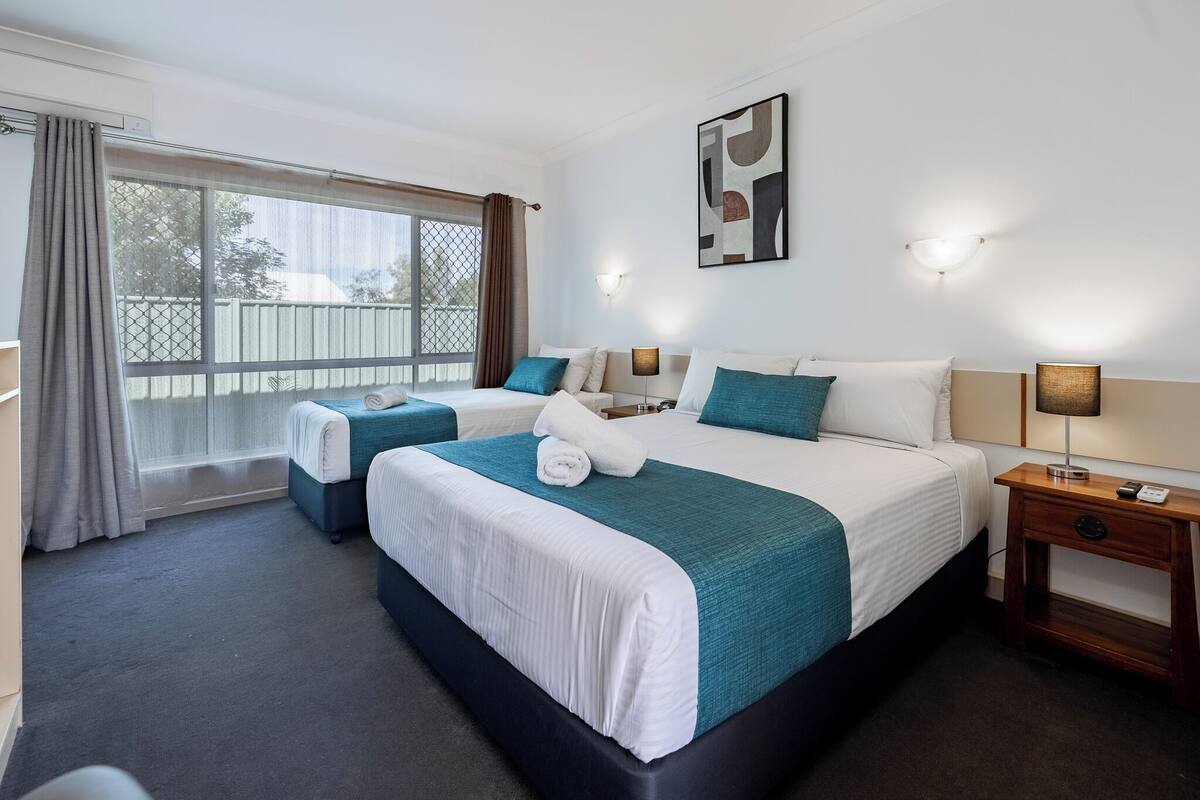 A hotel room with two beds, a large window with curtains, a wall-mounted artwork, and a bedside table with a lamp.