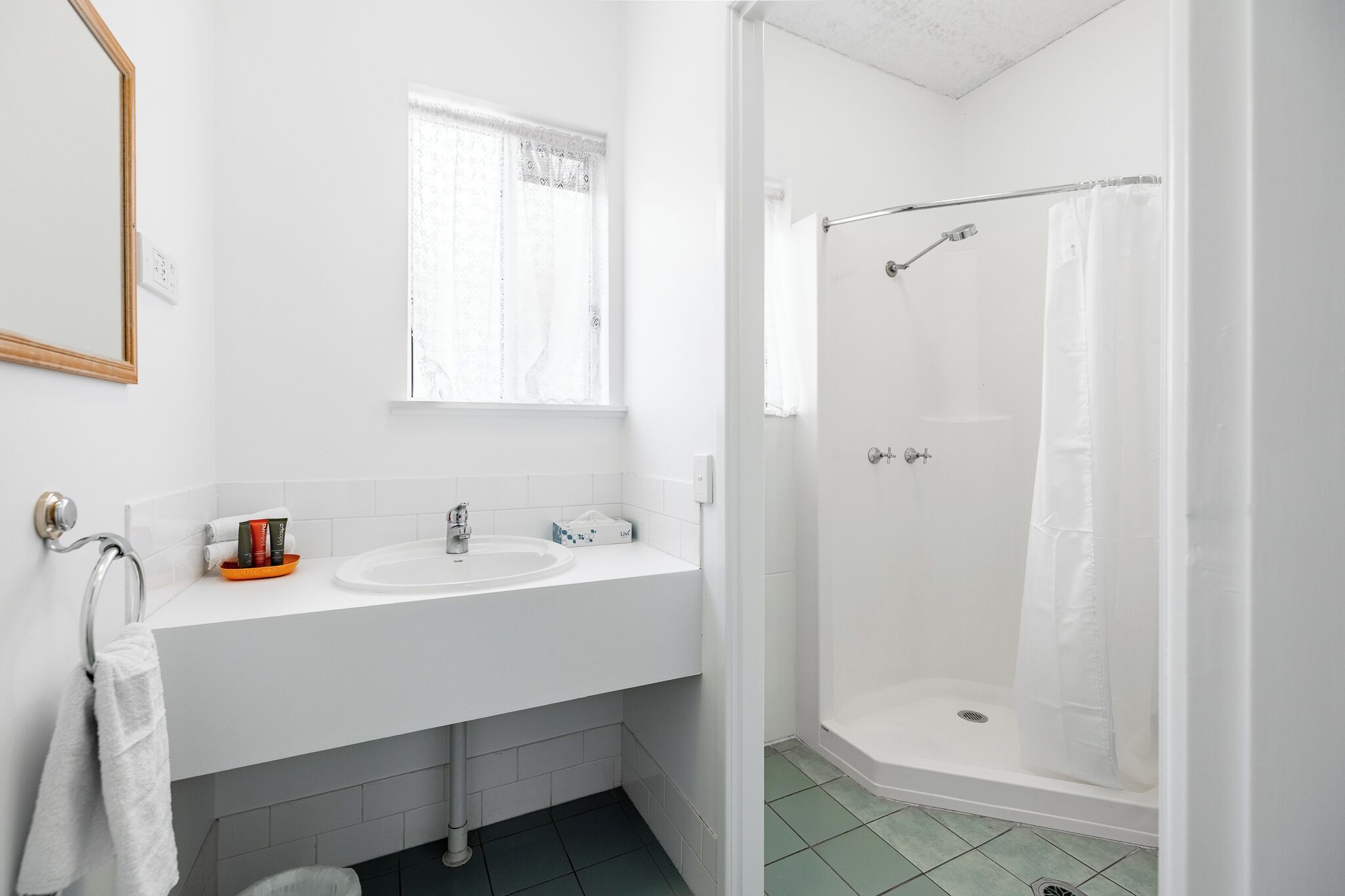 A bathroom with a white sink, a mirror, a window with lace curtains, and a shower with a glass door.