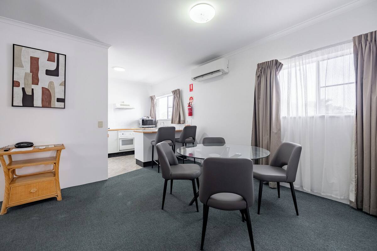 A modern hotel room with a round glass table, four grey chairs, a small wooden side table, a wall-mounted air conditioner, a fire extinguisher, and a window with sheer curtains.