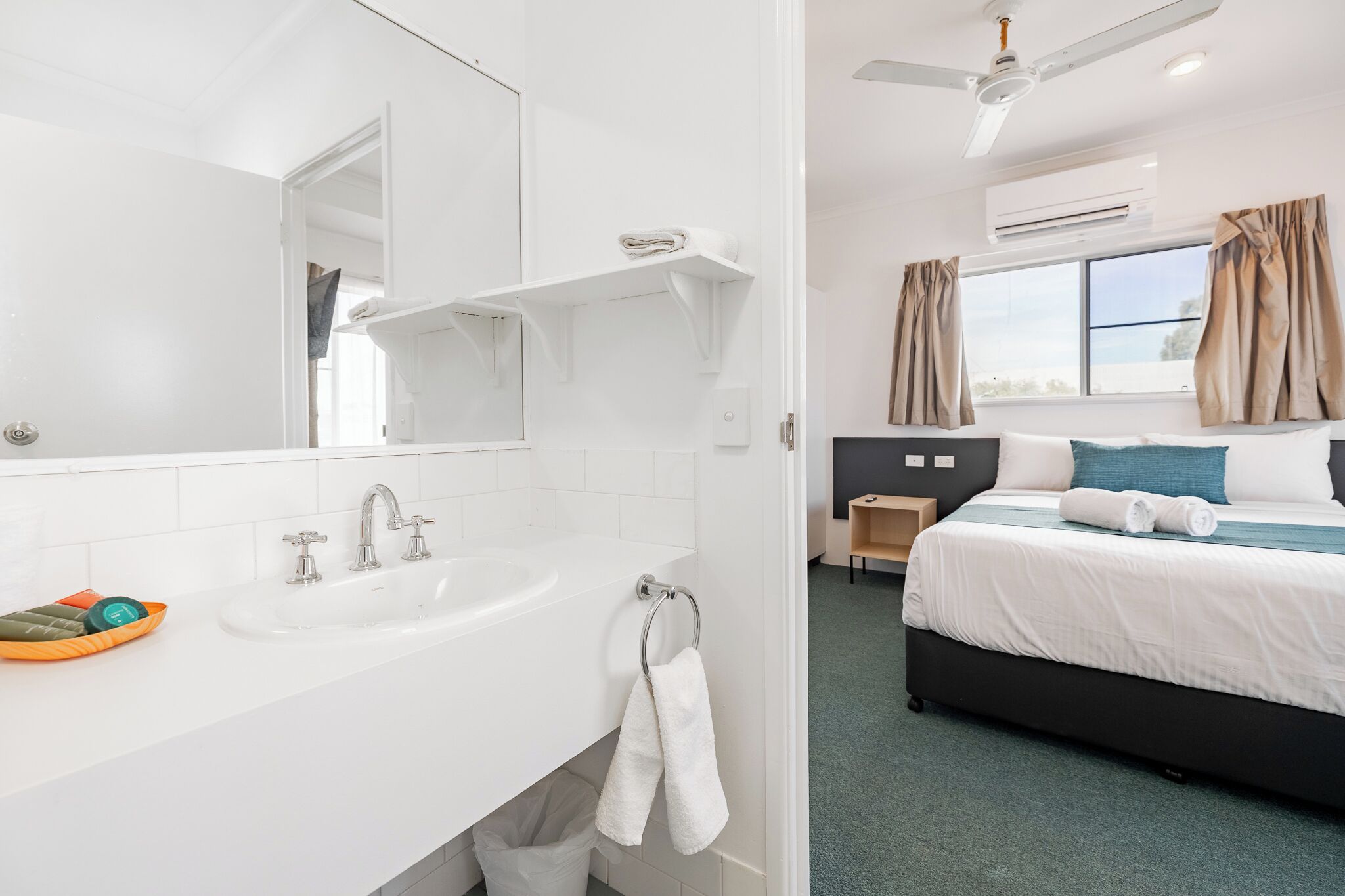 Une chambre d’hôtel avec un lavabo blanc, un miroir et un lit avec un oreiller bleu.