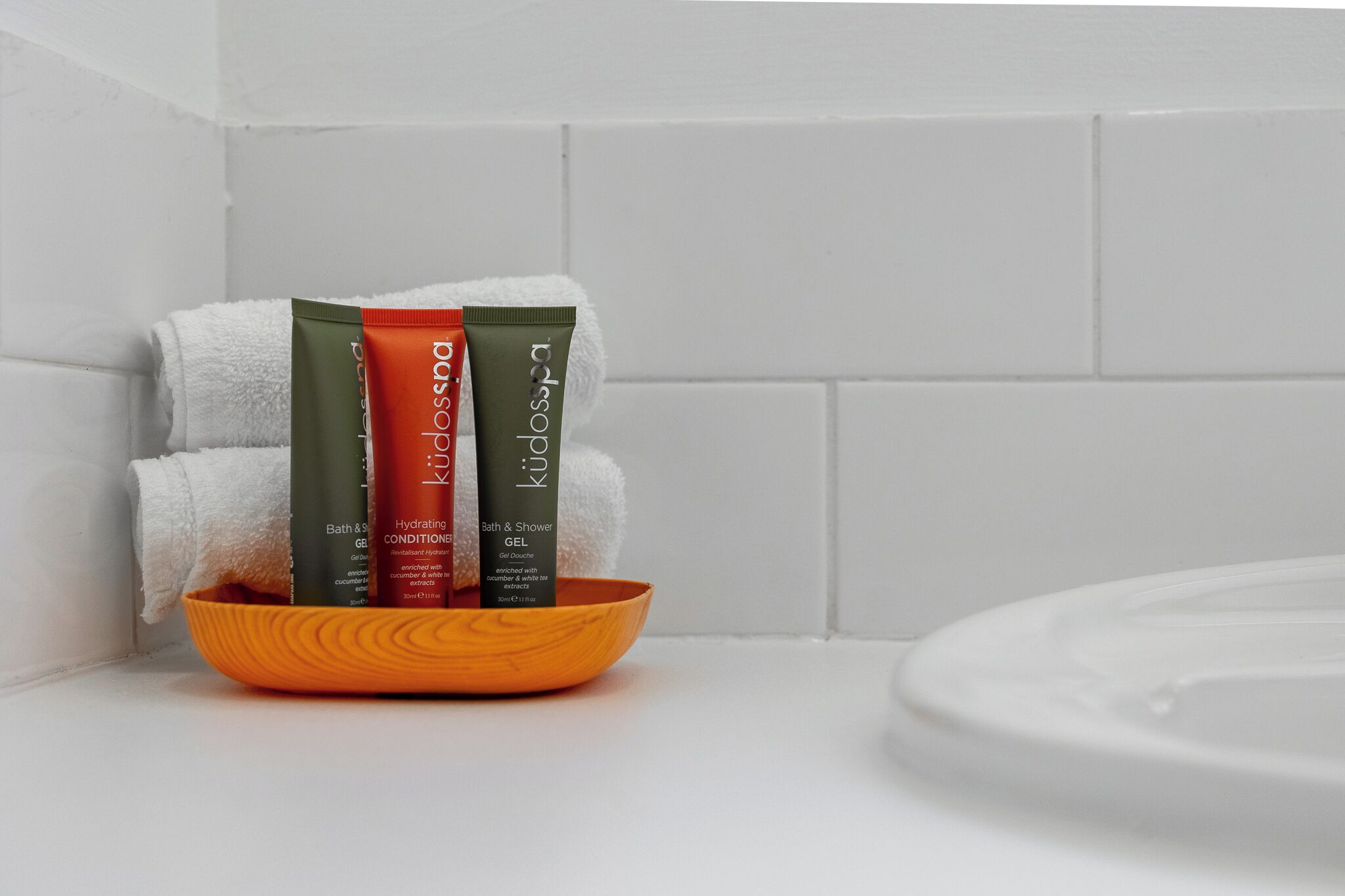 A bathroom with a white tiled wall, a white towel, and three bath products in a wooden bowl.