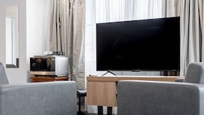 A hotel room with a flat-screen TV on a wooden stand, a microwave, a wooden desk, and grey upholstered chairs.