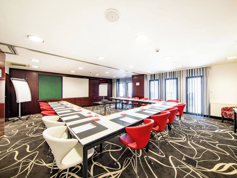 A conference room with a long table, red and white chairs, a whiteboard, and a green screen.