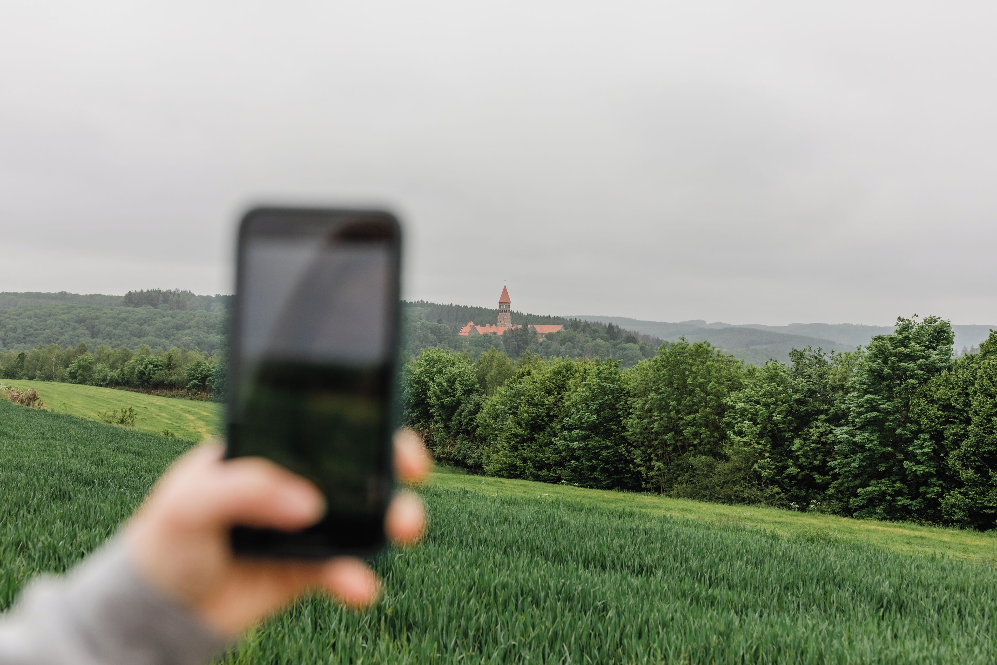 A hand holding a smartphone with a view of a green field and a distant building with a red roof.