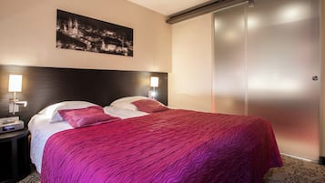 Hotellihuone, jossa on sänky, kaksi yöpöytävalaisinta, yöpöytä, seinälle ripustettu taulu ja liukuovilla varustettu vaatekaappi.