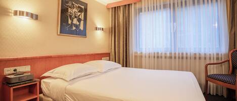 Habitación de hotel con cama, una silla, una mesita de noche con radio y un cuadro en la pared.