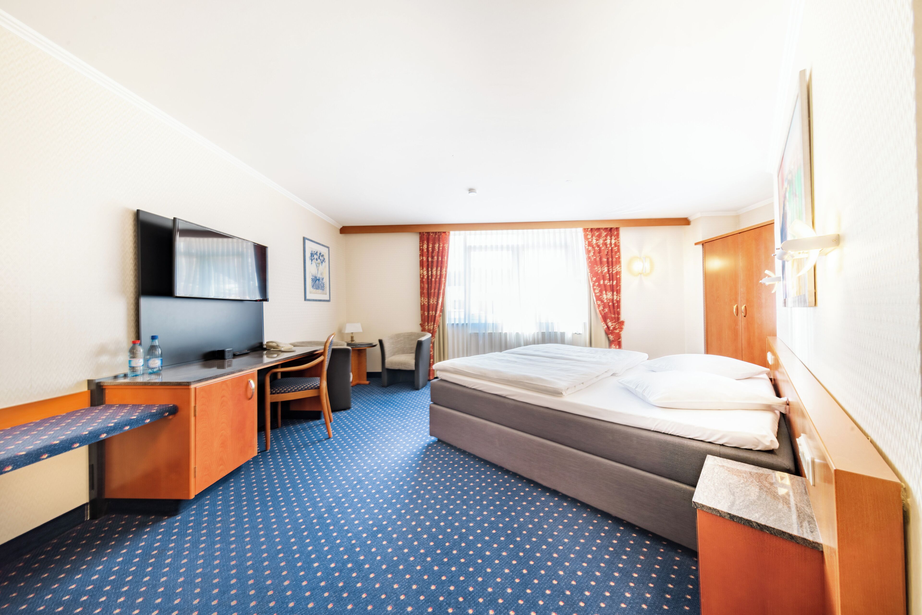 Ein Hotelzimmer mit Bett, Schreibtisch, Stuhl, Fernseher und einem Fenster mit Vorhängen.