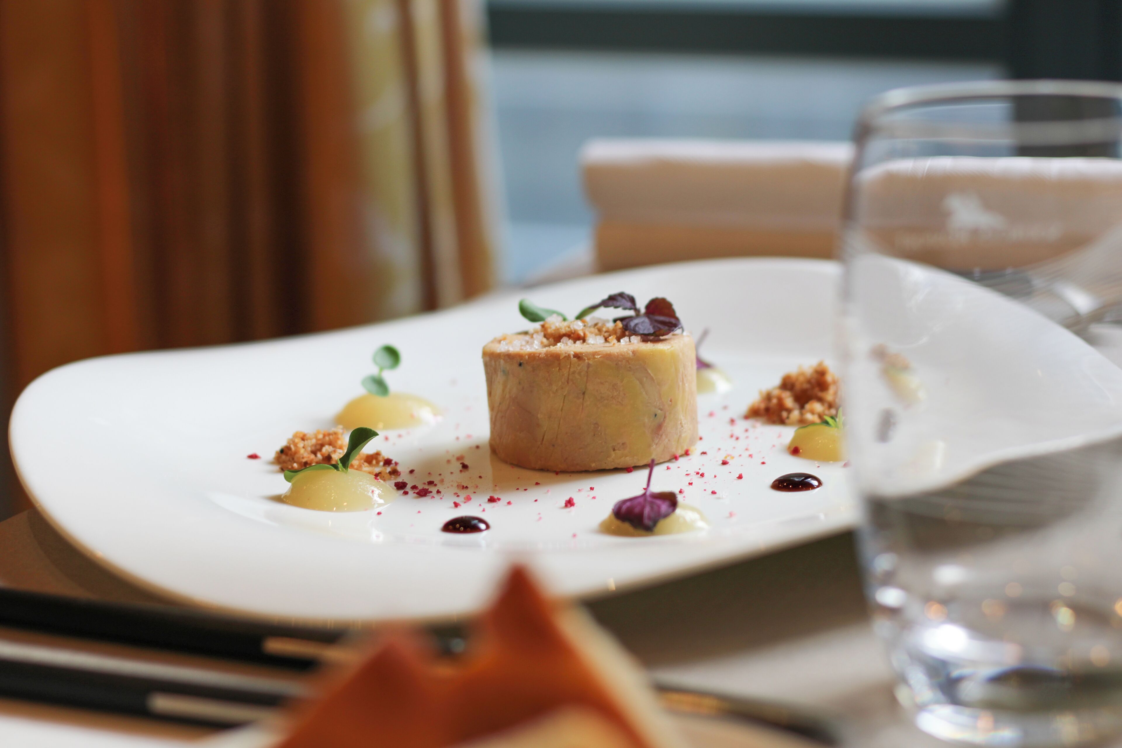 A dessert on a white plate with garnishes and a glass of water on the table.