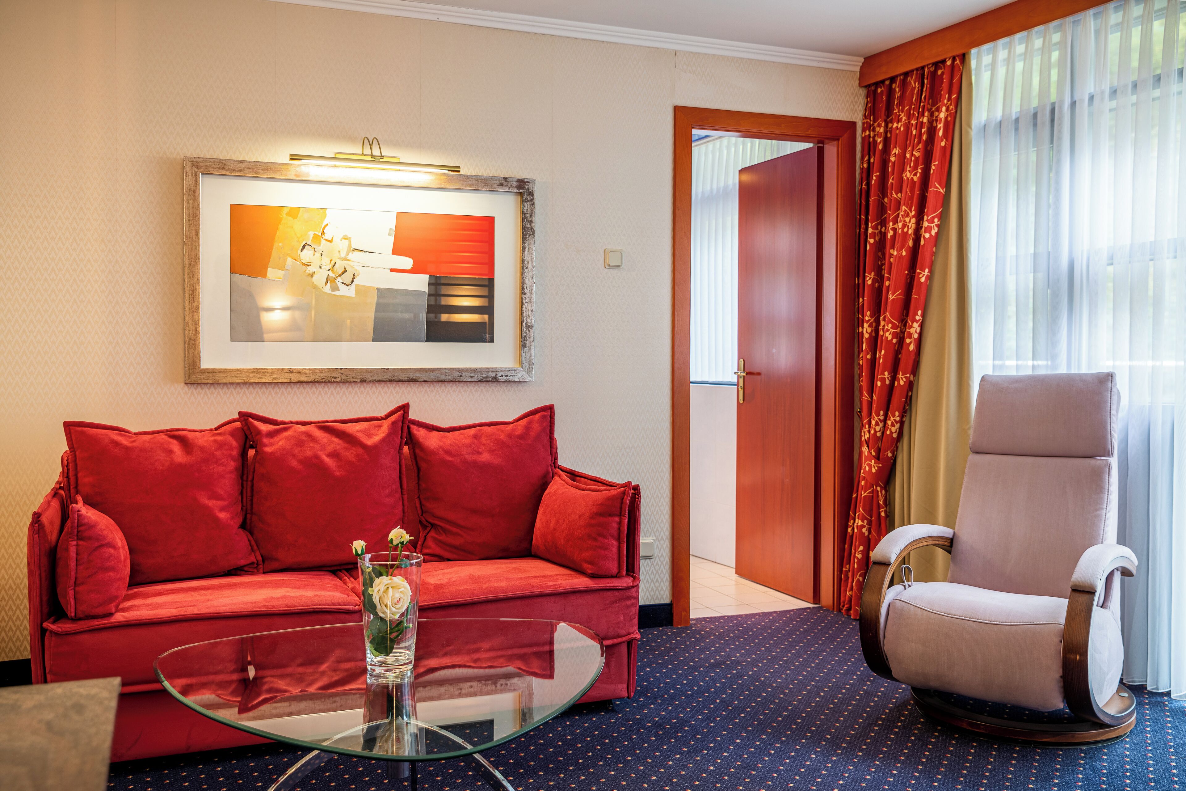 Ein Hotelzimmer mit einem roten Sofa, einem Glastisch, einem weißen Sessel und einem gerahmten abstrakten Gemälde an der Wand.