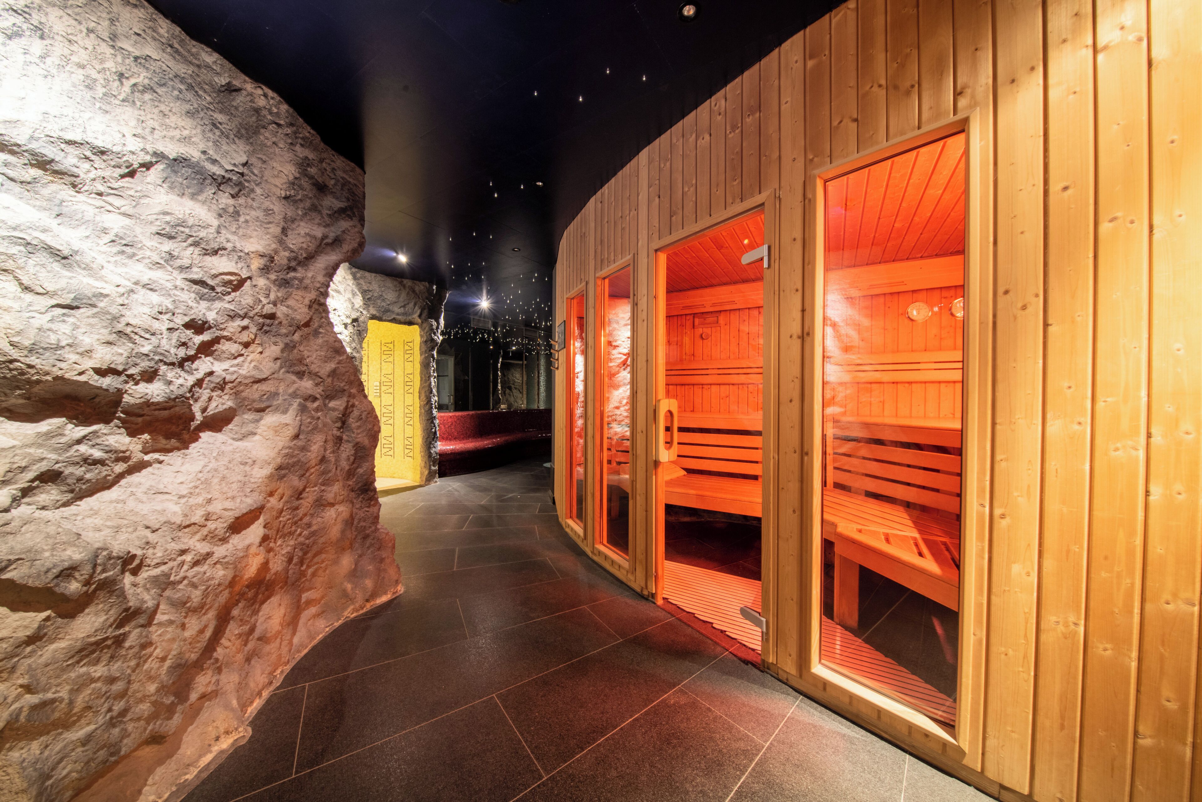 Un pasillo con paredes y bancos de madera, que conduce a una zona de sauna con bancos rojos y una pared de piedra en un lateral.