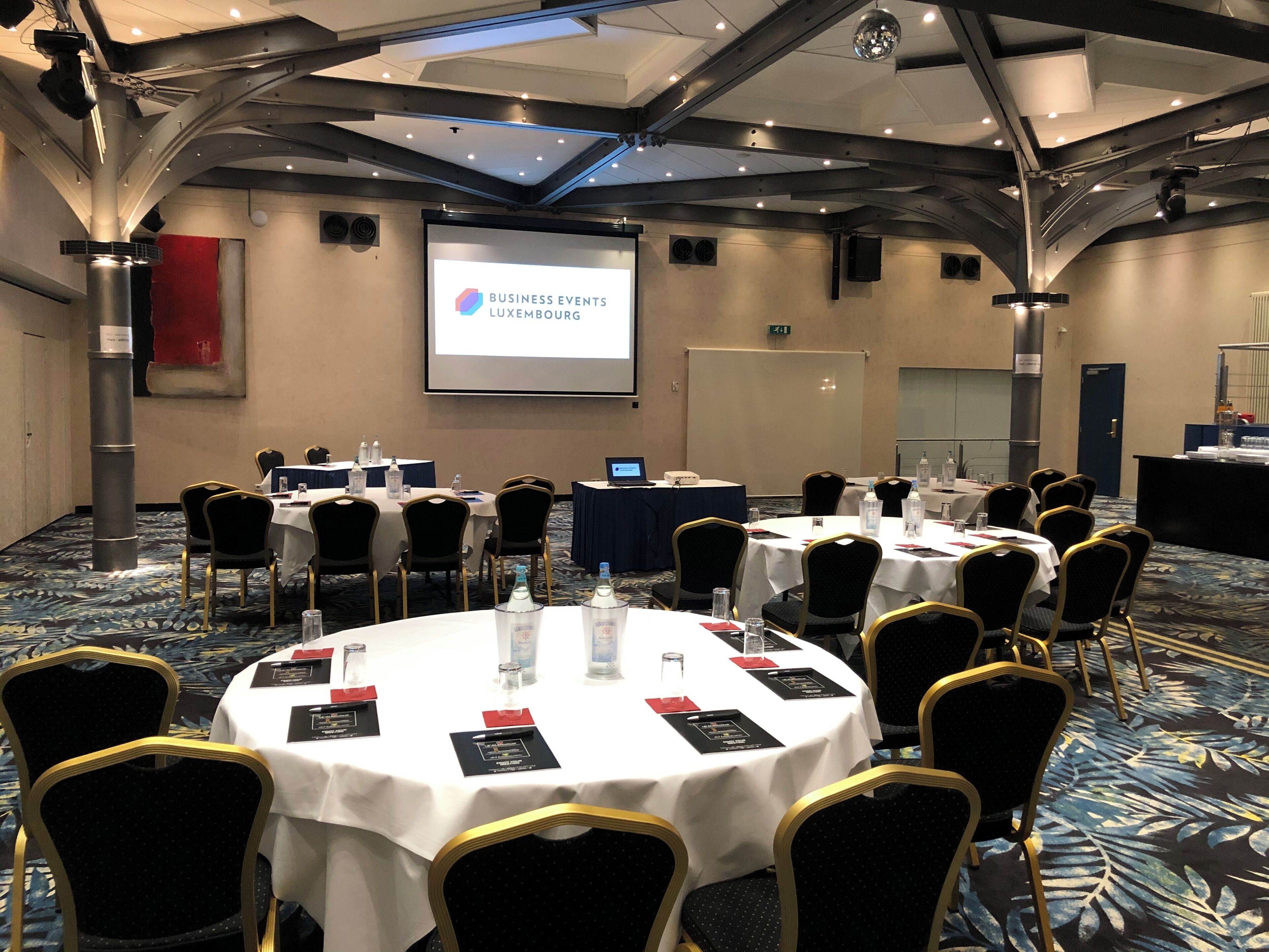 Ein Konferenzraum mit runden Tischen, schwarzen Stühlen mit goldenen Akzenten und einer Projektionsleinwand, die "BUSINESS EVENTS LUXEMBOURG" zeigt.