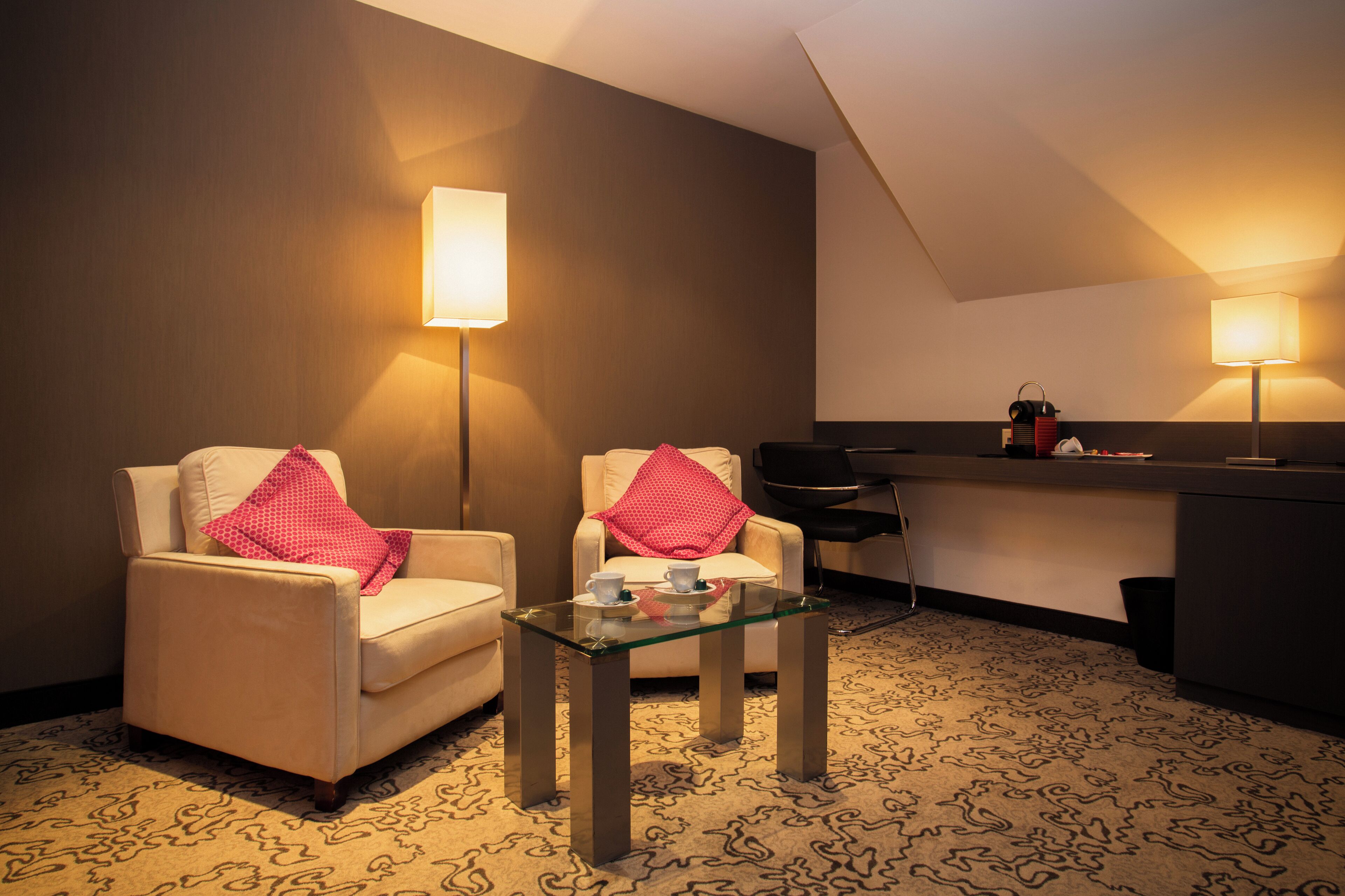 Une chambre d’hôtel avec deux fauteuils, une table basse en verre, un minibar et deux lampes fixées au mur.