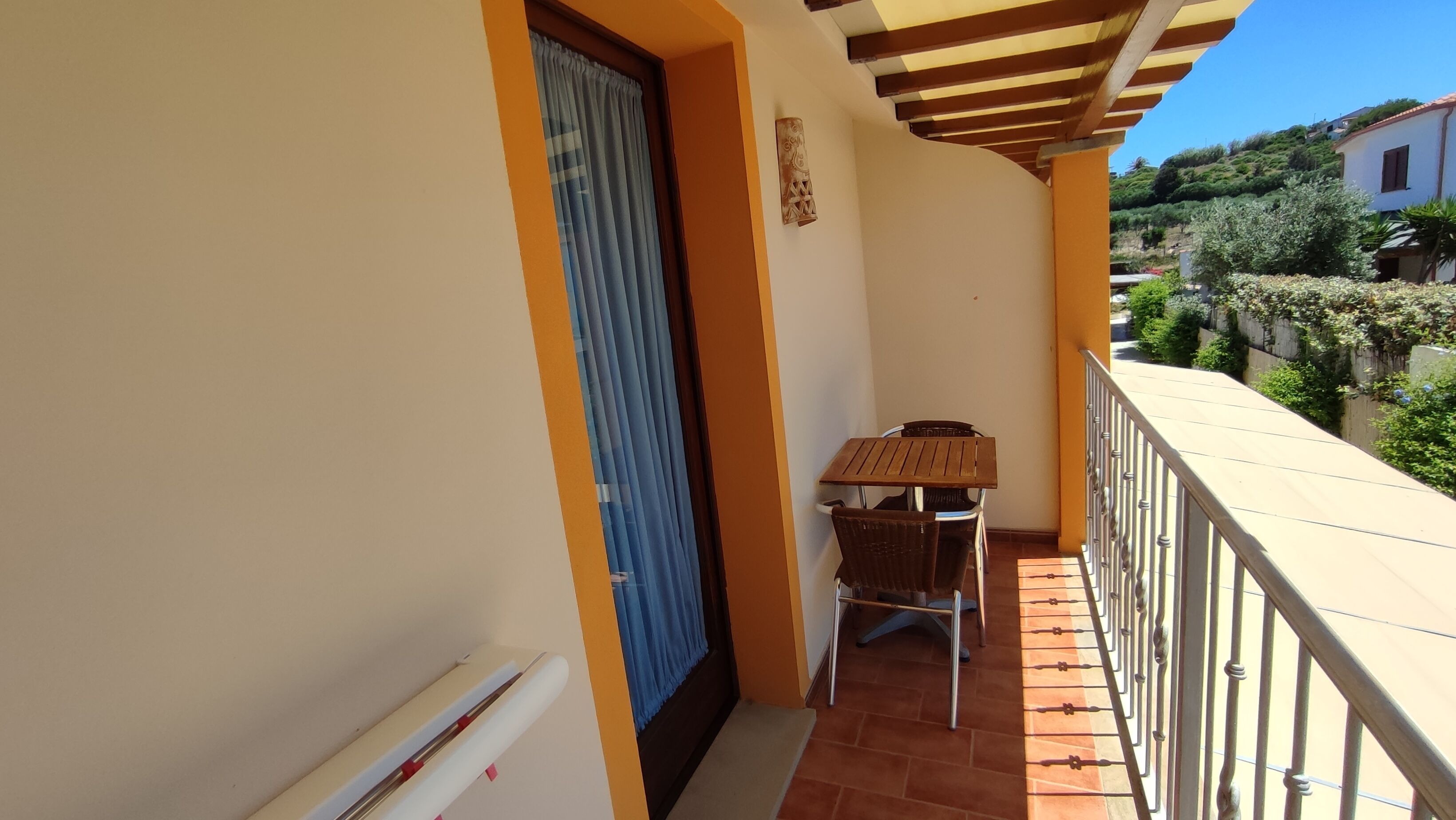 A balcony with a wooden table and chairs, a blue door, and a view of greenery and houses.