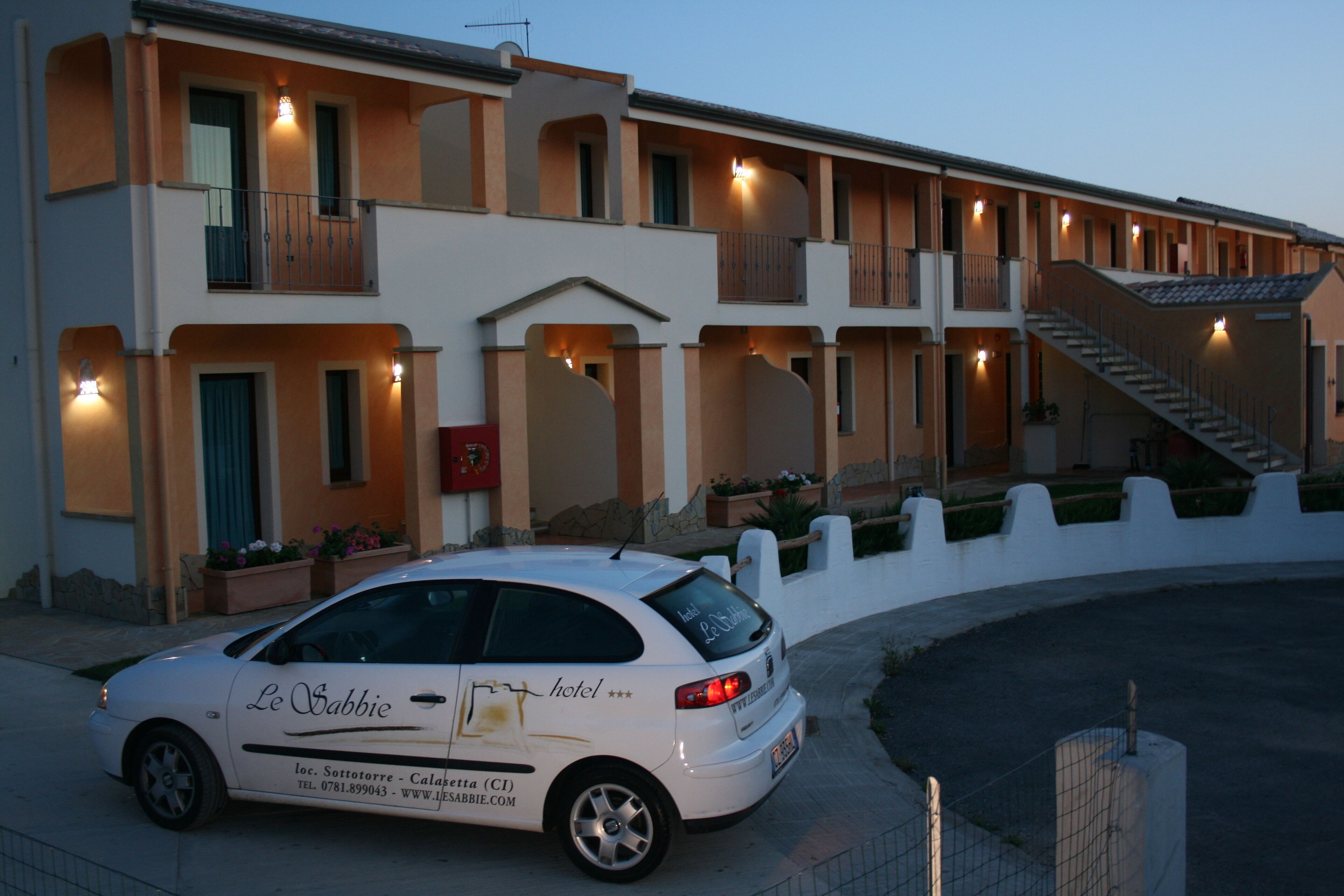 A hotel with a white facade, multiple balconies, and a car parked in front.