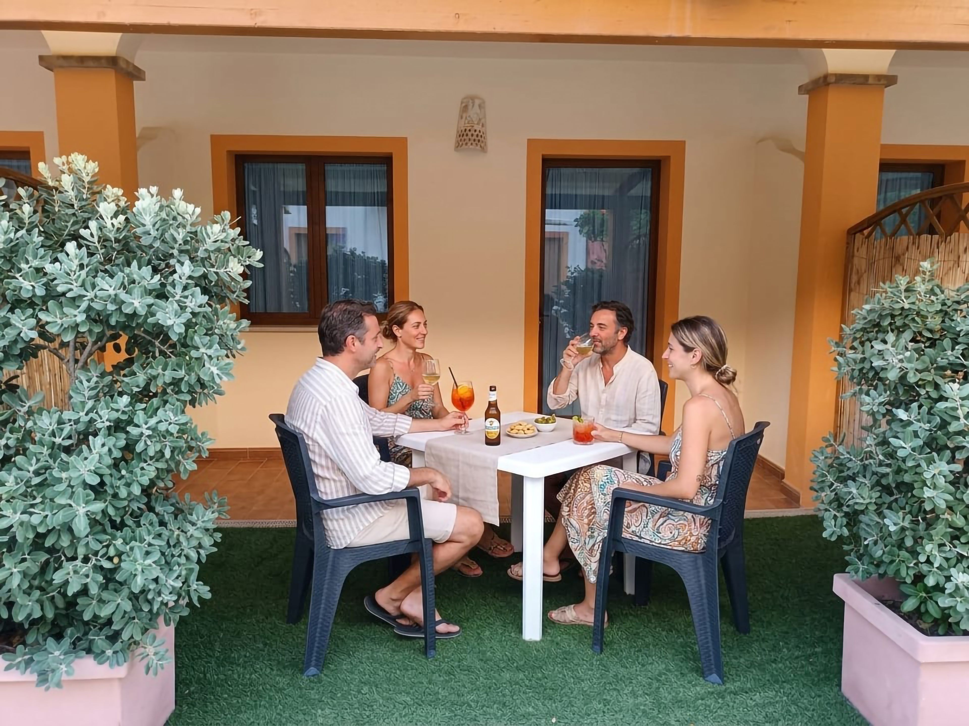 A group of people seated at an outdoor table with a drink and snacks, surrounded by potted plants and a building with windows.