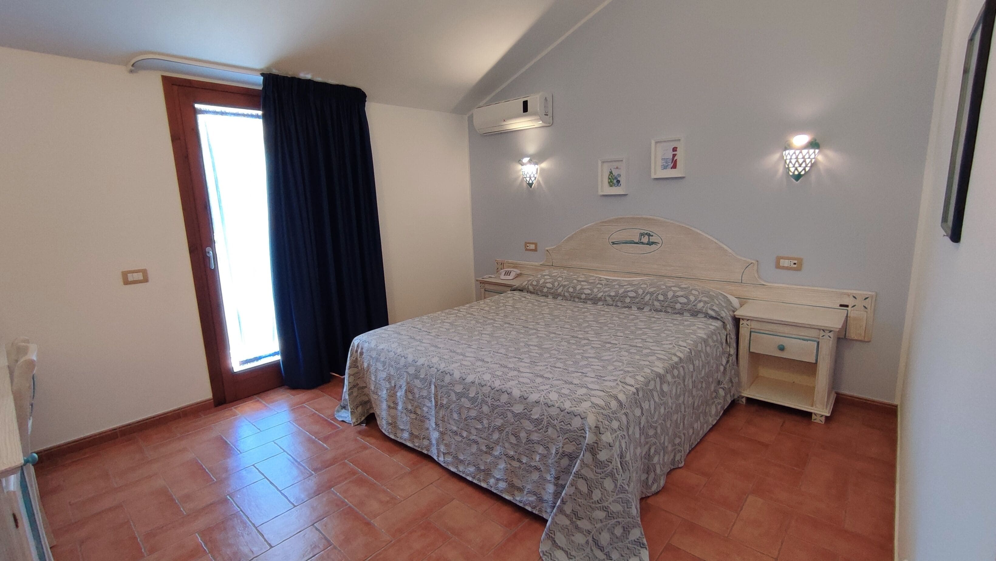Quarto com cama, mesas de cabeceira, um criado-mudo, janela com cortinas e luminárias na parede.
