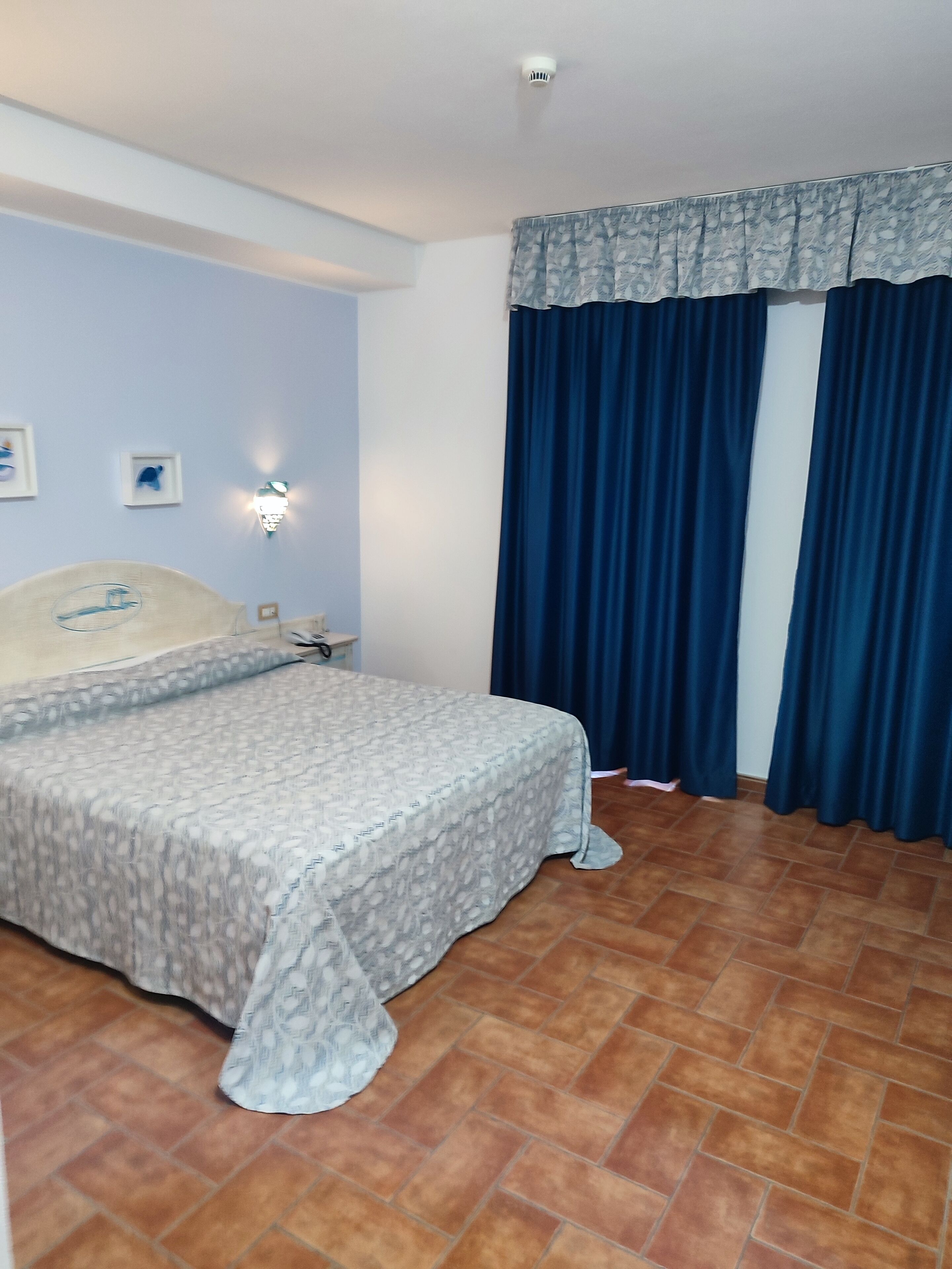 A bedroom with a bed, a nightstand, a wall-mounted lamp, and a window with curtains.