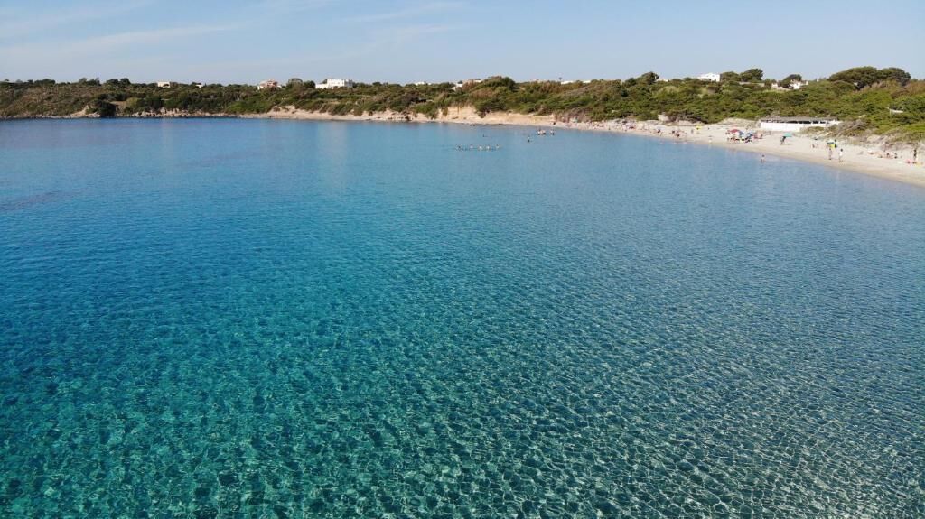 Una playa costera con agua azul cristalina, orilla de arena y edificios al fondo.