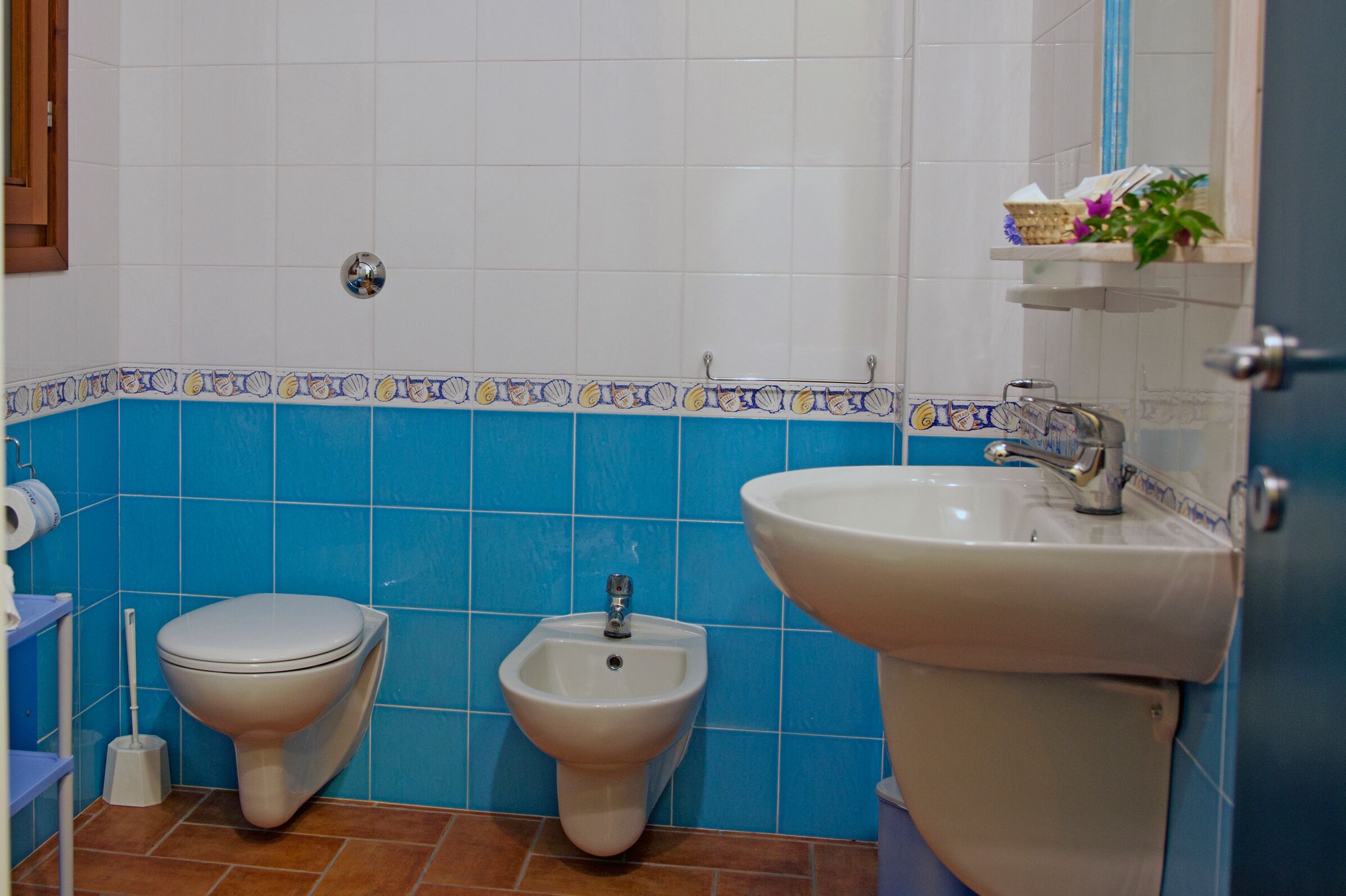 A bathroom with a toilet, bidet, and sink, blue and white tiled walls, and a small shelf with toiletries.