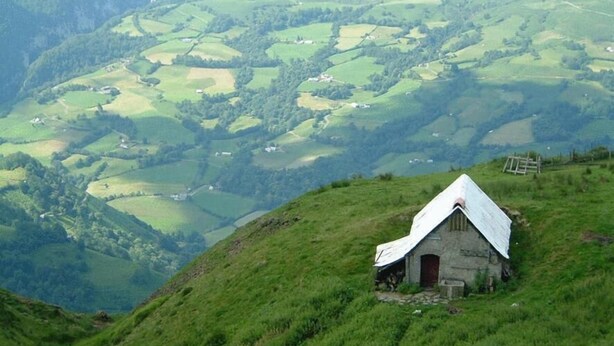 Una pequeña casa de piedra con techo blanco sobre una colina cubierta de hierba, rodeada de campos verdes ondulantes y un valle.