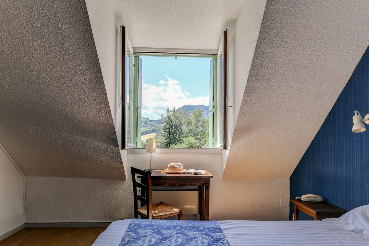 A bedroom with a bed, a desk with a lamp, a chair, and a view of the outdoors through a window.