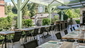 Un área de comedor al aire libre con mesas y sillas, un pasillo cubierto y un edificio de ladrillo al fondo.
