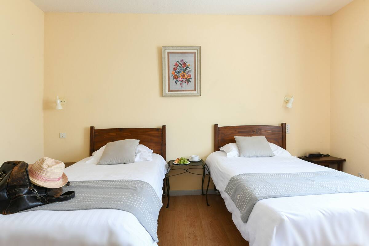 A hotel room with two beds, a nightstand with a lamp, a framed floral artwork on the wall, and a suitcase on the floor.