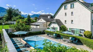 Zona de piscina con sillones reclinables y sombrillas, un edificio grande con techo inclinado y montañas al fondo.