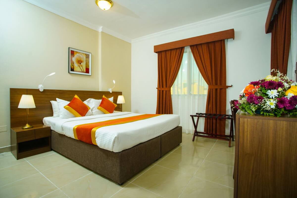 A hotel room with a large bed, wooden headboard, two bedside tables with lamps, a desk with a chair, and a window with curtains.