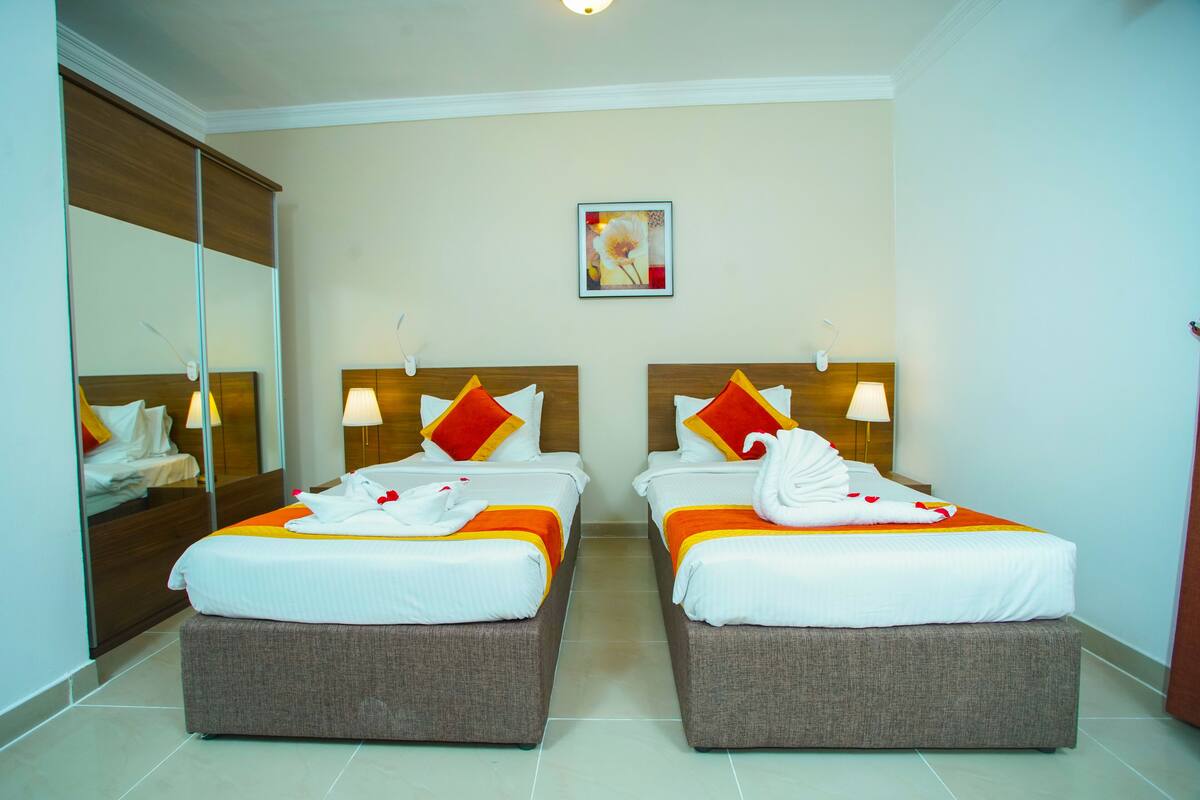 A hotel room with two beds, each with a wooden headboard, white bedding, and red and yellow pillows. There is a large mirror on one wall and a framed picture above the beds.