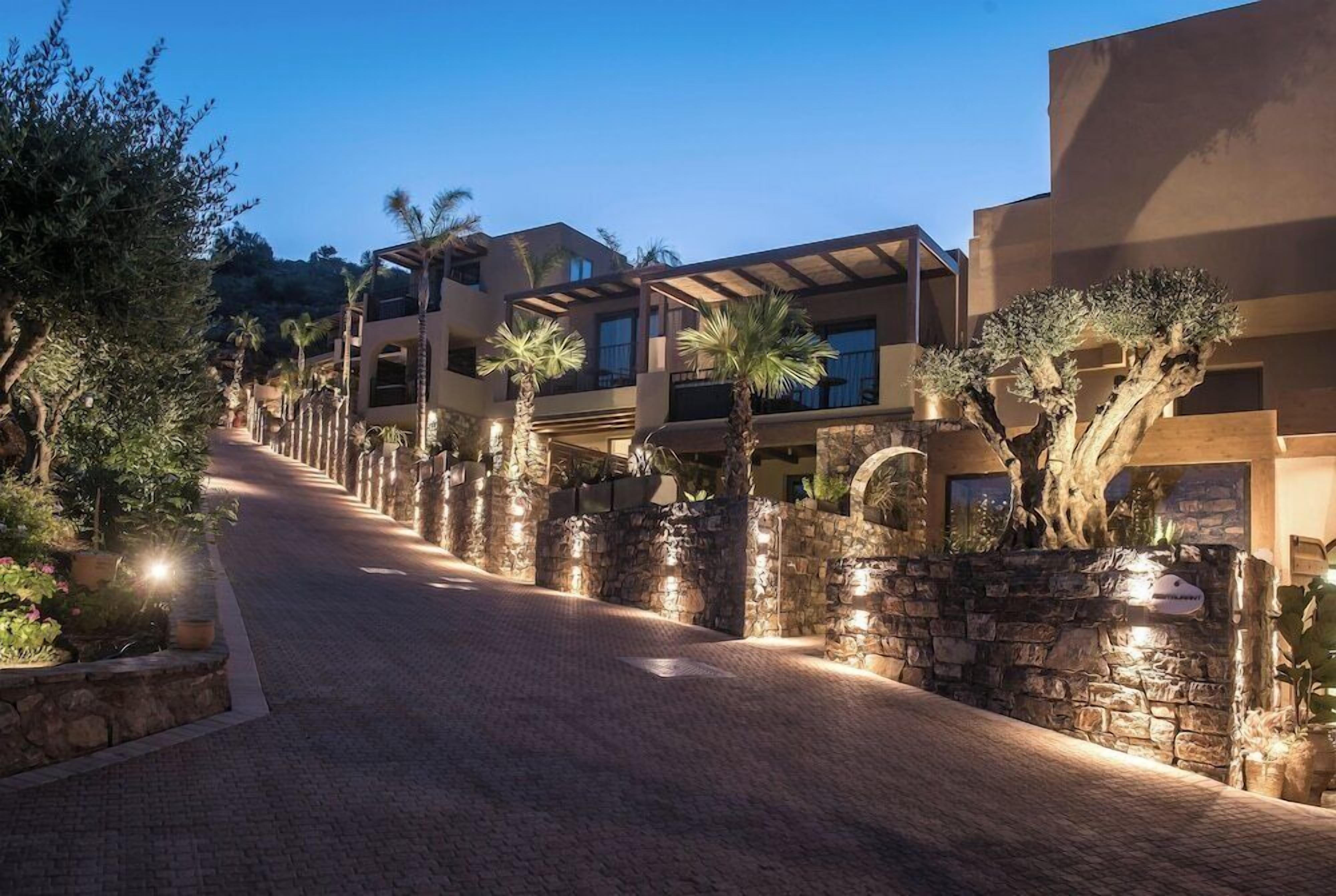 Un hotel de varios pisos con un camino pavimentado, muros de piedra y palmeras.