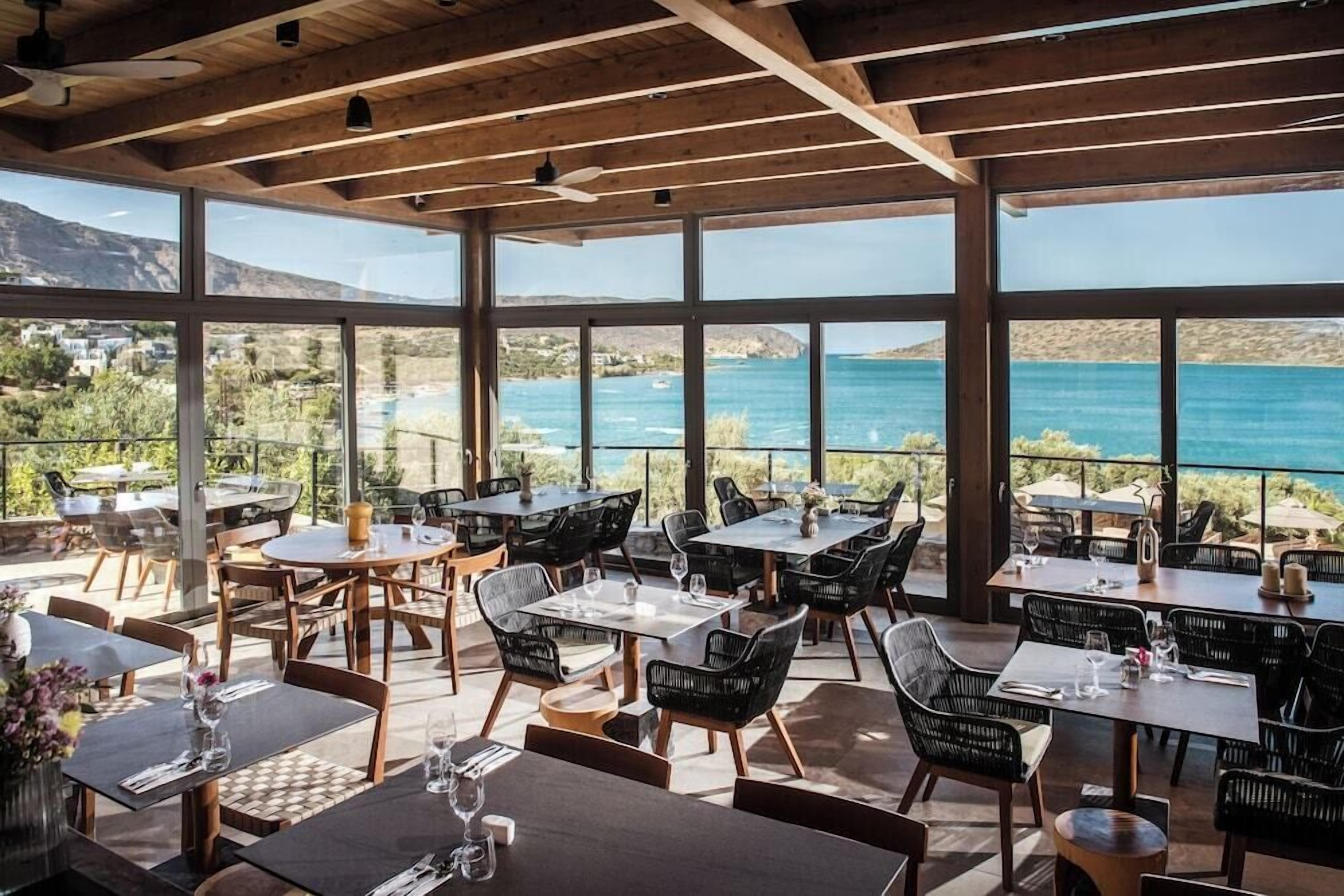 Un área de comedor al aire libre con mesas y sillas, con vista a un cuerpo de agua y colinas.
