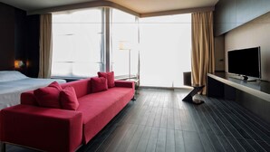 A modern hotel room with a red sofa, a flat-screen TV, a bed, and large windows with curtains. - Riviera Golf Resort (San Giovanni In Marignano)